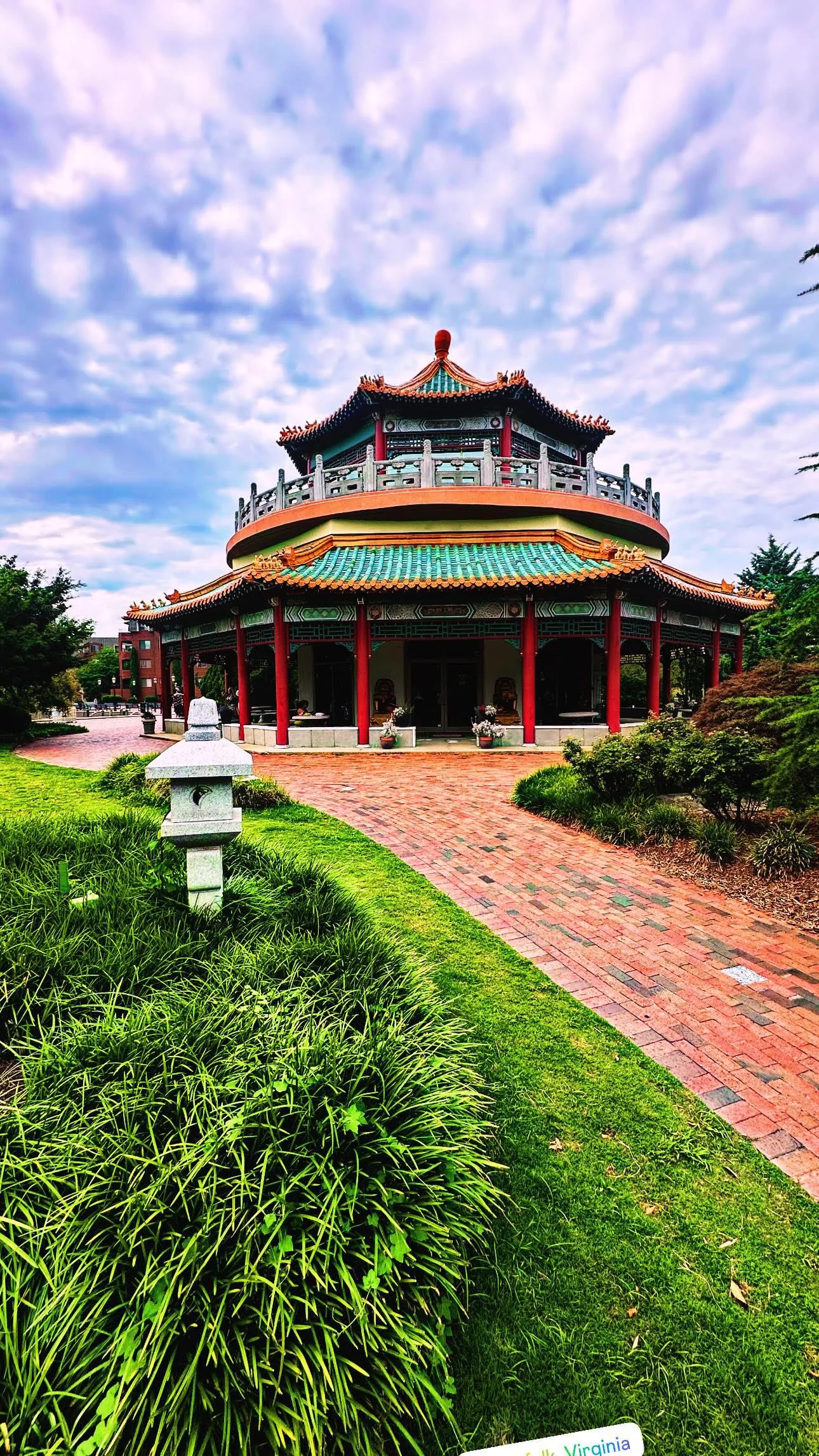 A traditional Chinese pavilion with a curved teal roof and red columns, surrounded by lush green grass and shrubs, with a red brick pathway leading to it, under a partly cloudy sky.