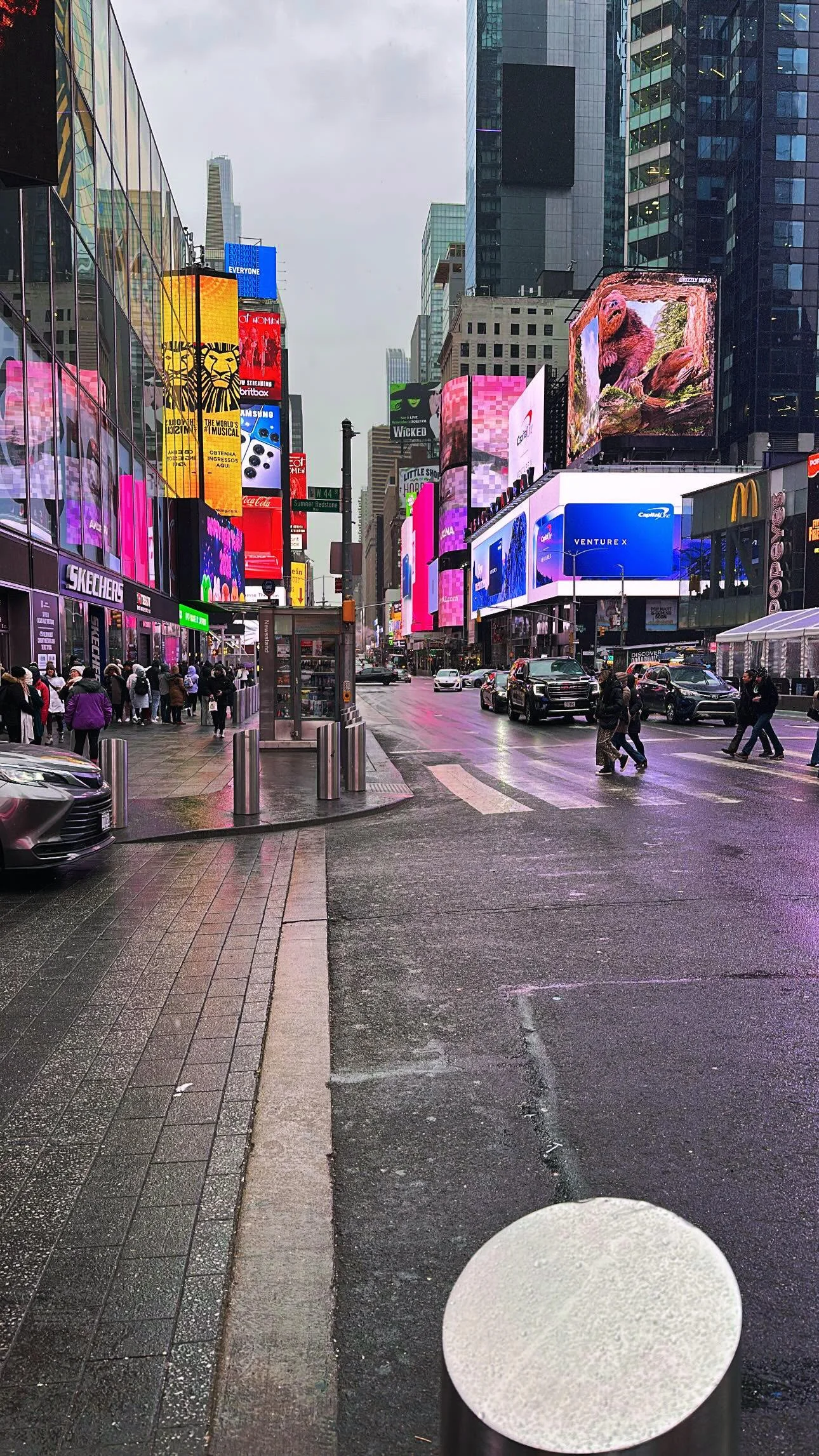 New York City Times Square with digital billboards, tall buildings, and pedestrians crossing the street on a wet day.