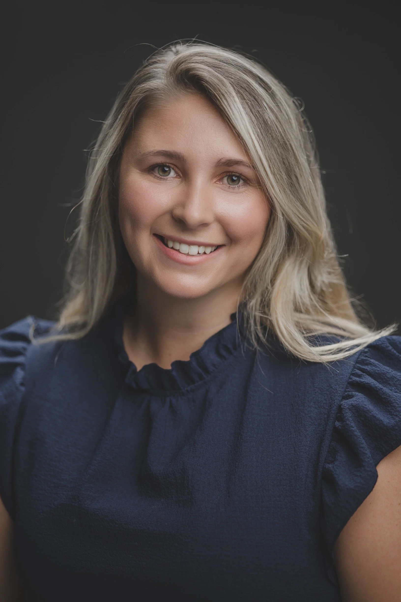 A woman with blonde hair and green eyes smiling at the camera, wearing a navy blue top with a ruffled collar.