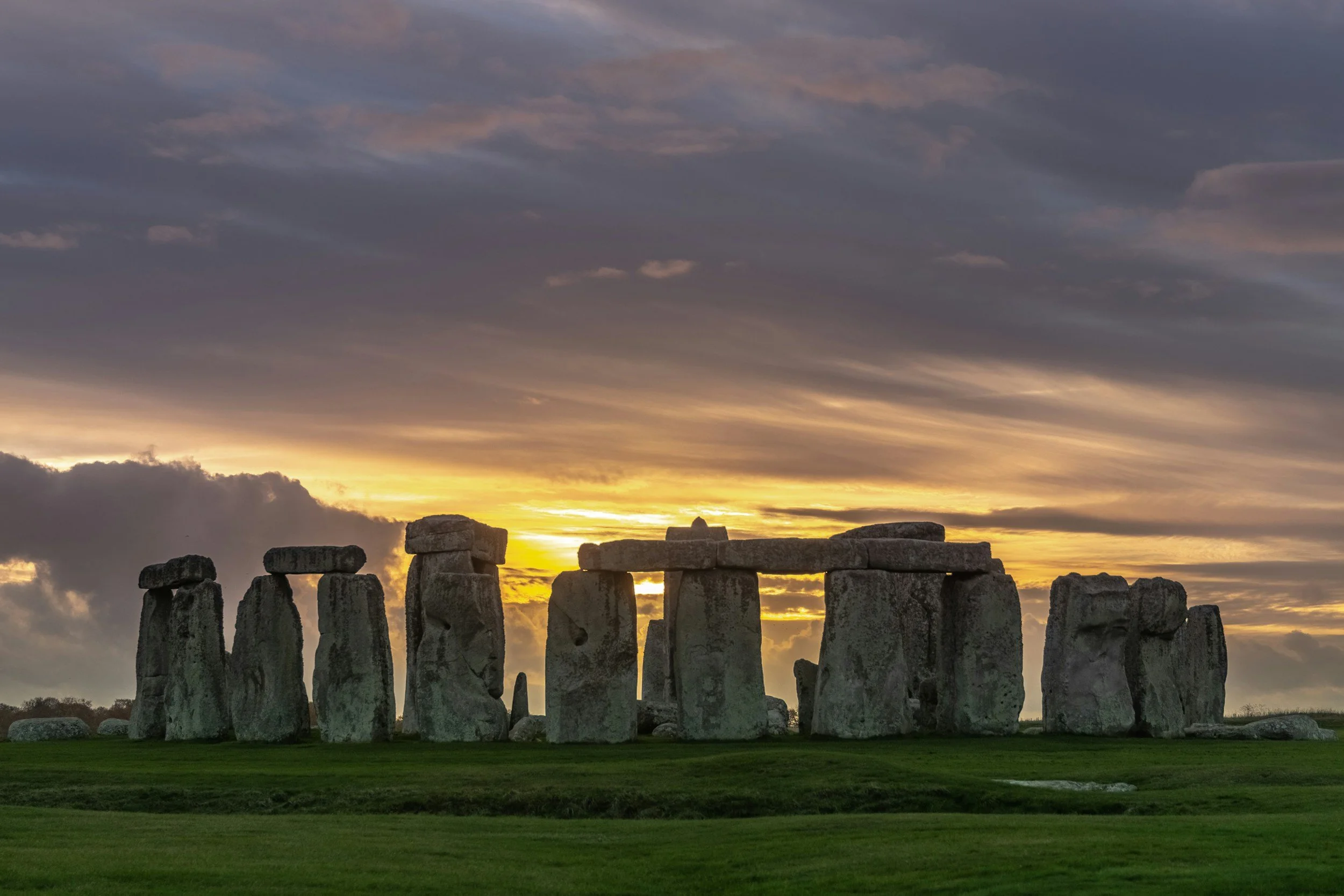Sunset view of Stonehenge with a dramatic cloudy sky overhead and lush green grass in the foreground.