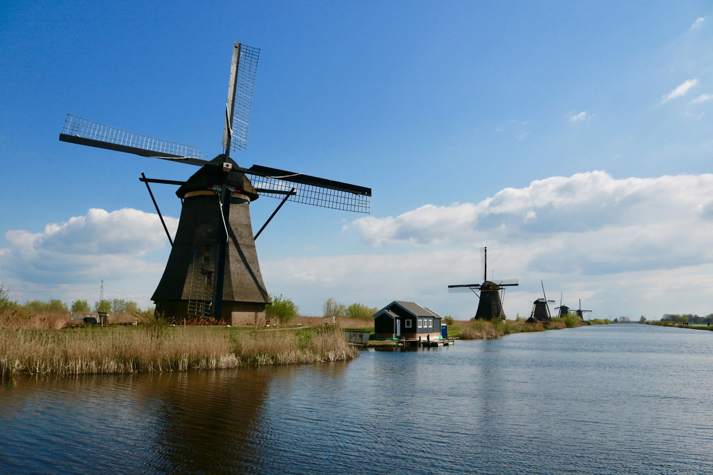 A scenic landscape with historic windmills along a river under a bright blue sky with scattered clouds.