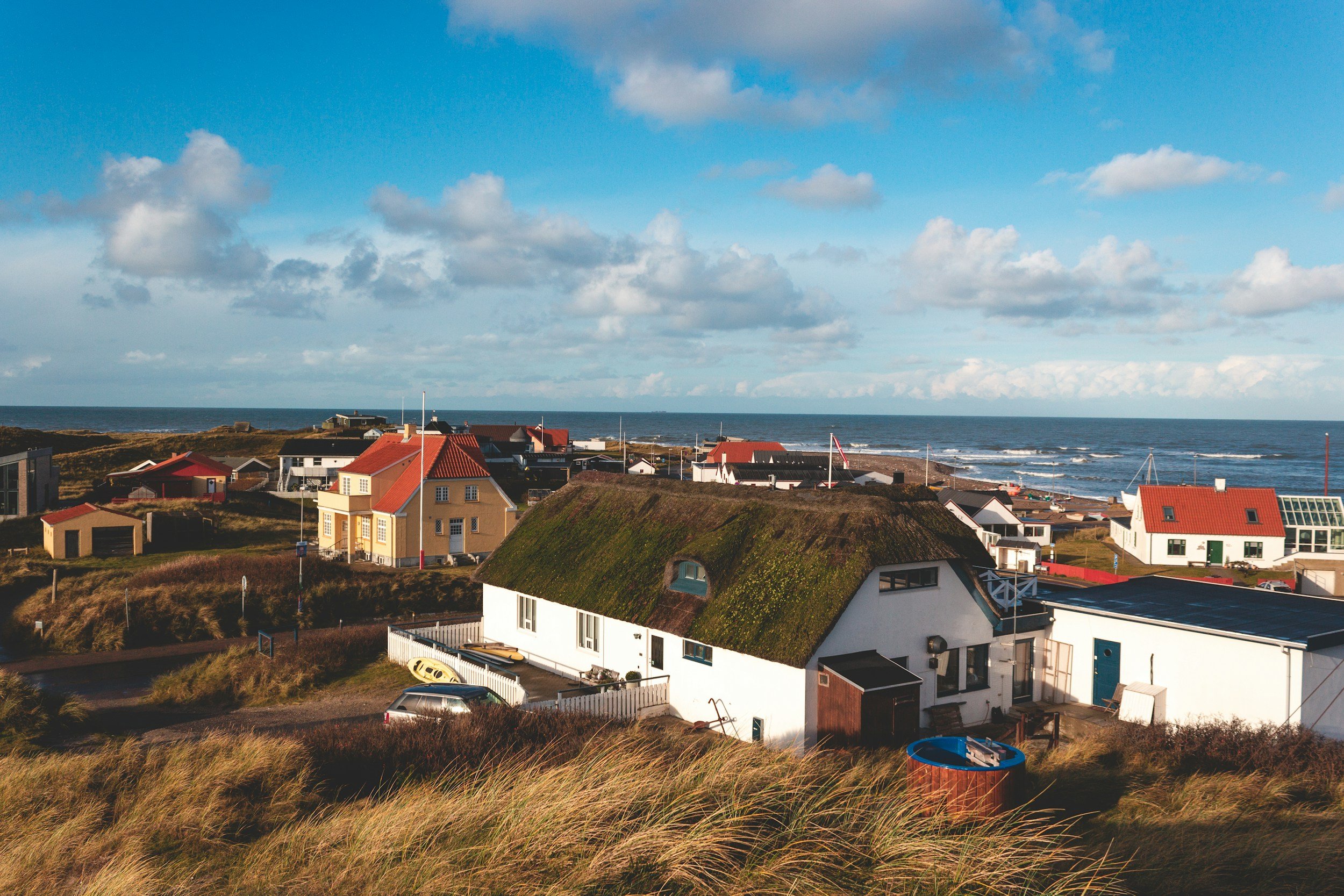 Coastal scene with houses, grassy dunes, and the ocean under partly cloudy sky.