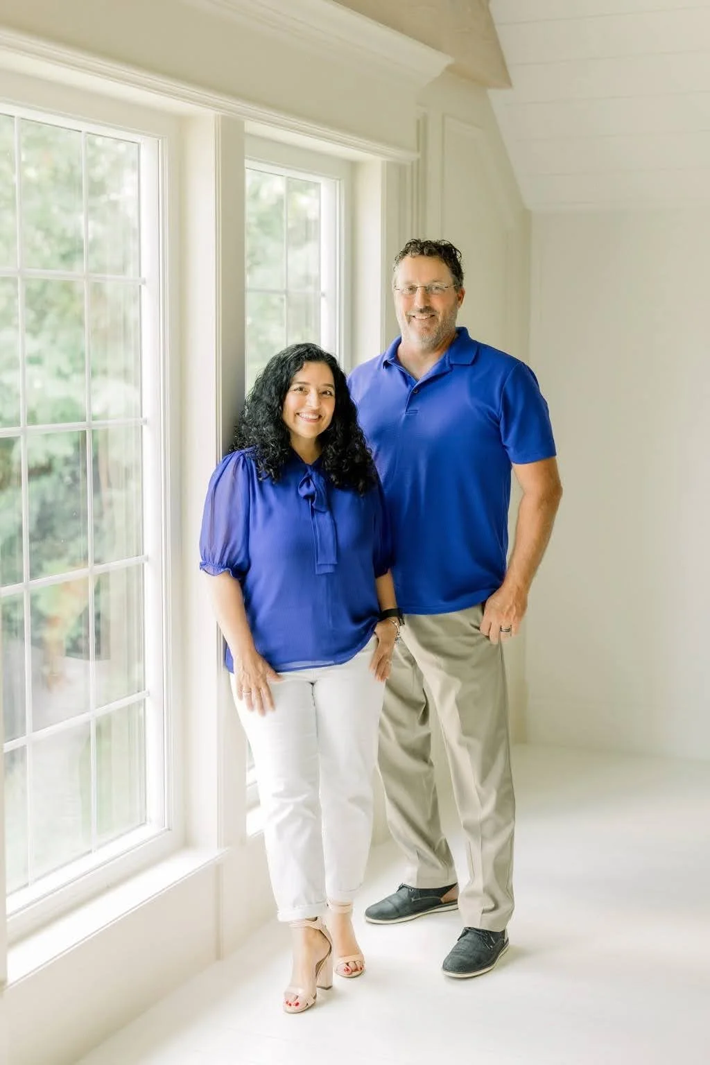 Jessica Fields is standing next to a window with a blue blouse, white pants, and nude heels her husband is with her in a matching blue shirt and khaki pants.