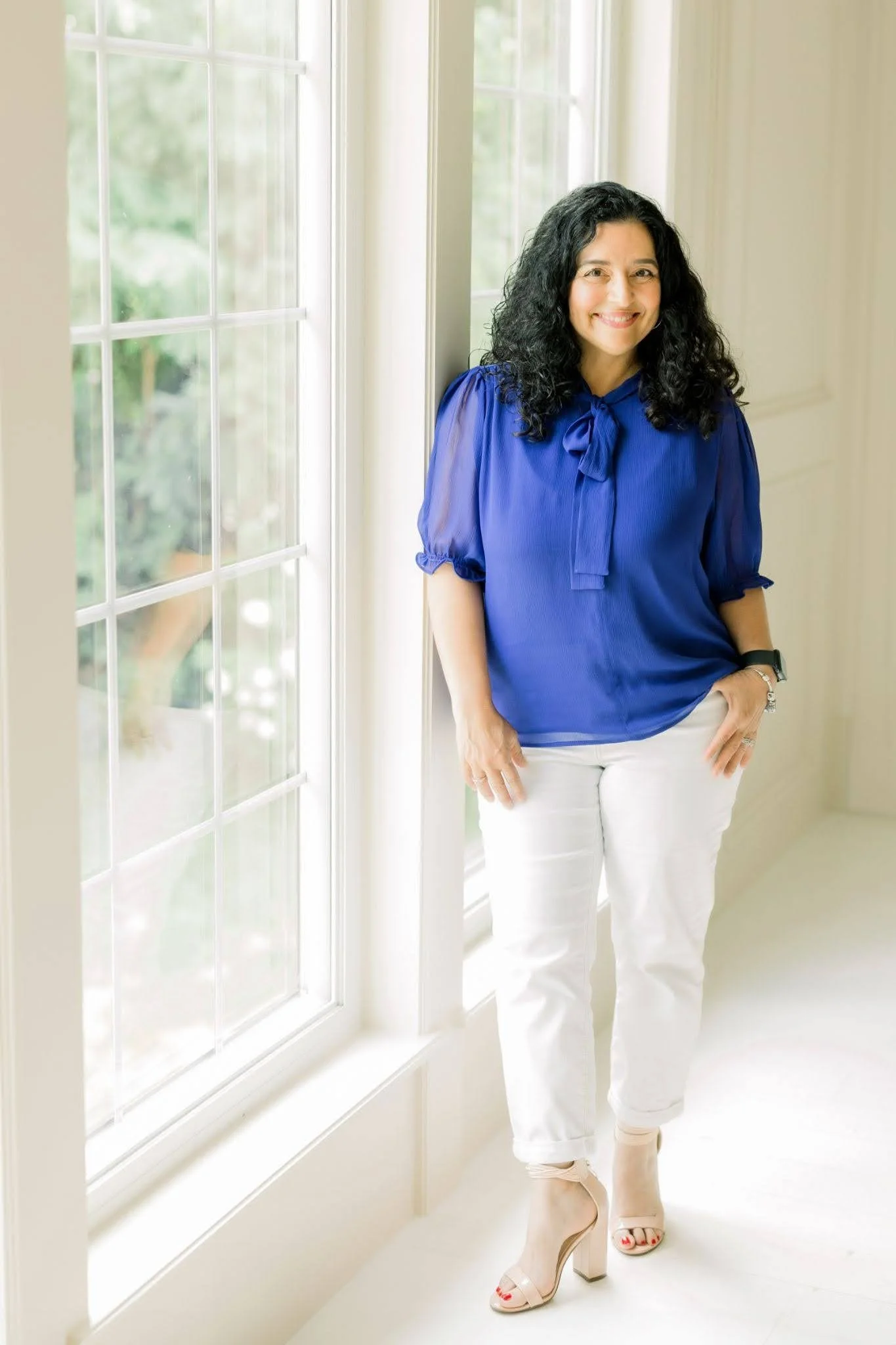 Jessica Fields is standing next to a window with a blue blouse, white pants, and nude heels.
