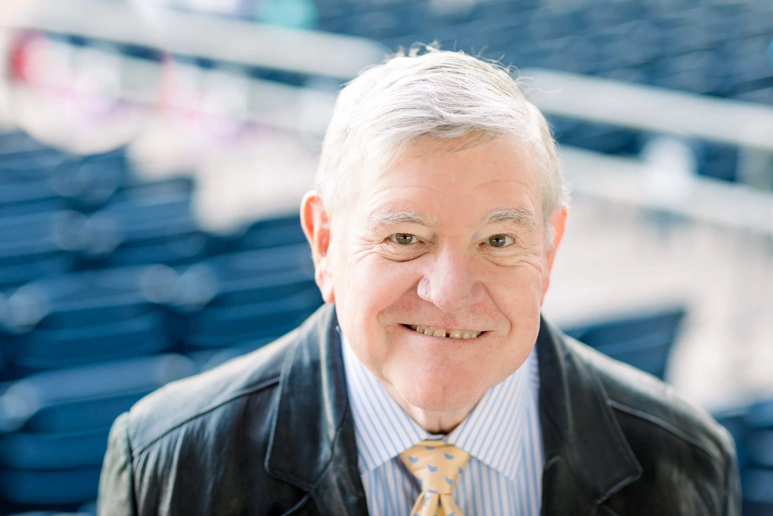 Close-up of an elderly man smiling, with short gray hair, wearing a dark blazer, light blue striped shirt, and yellow tie with blue stars, set against a blurred blue background.