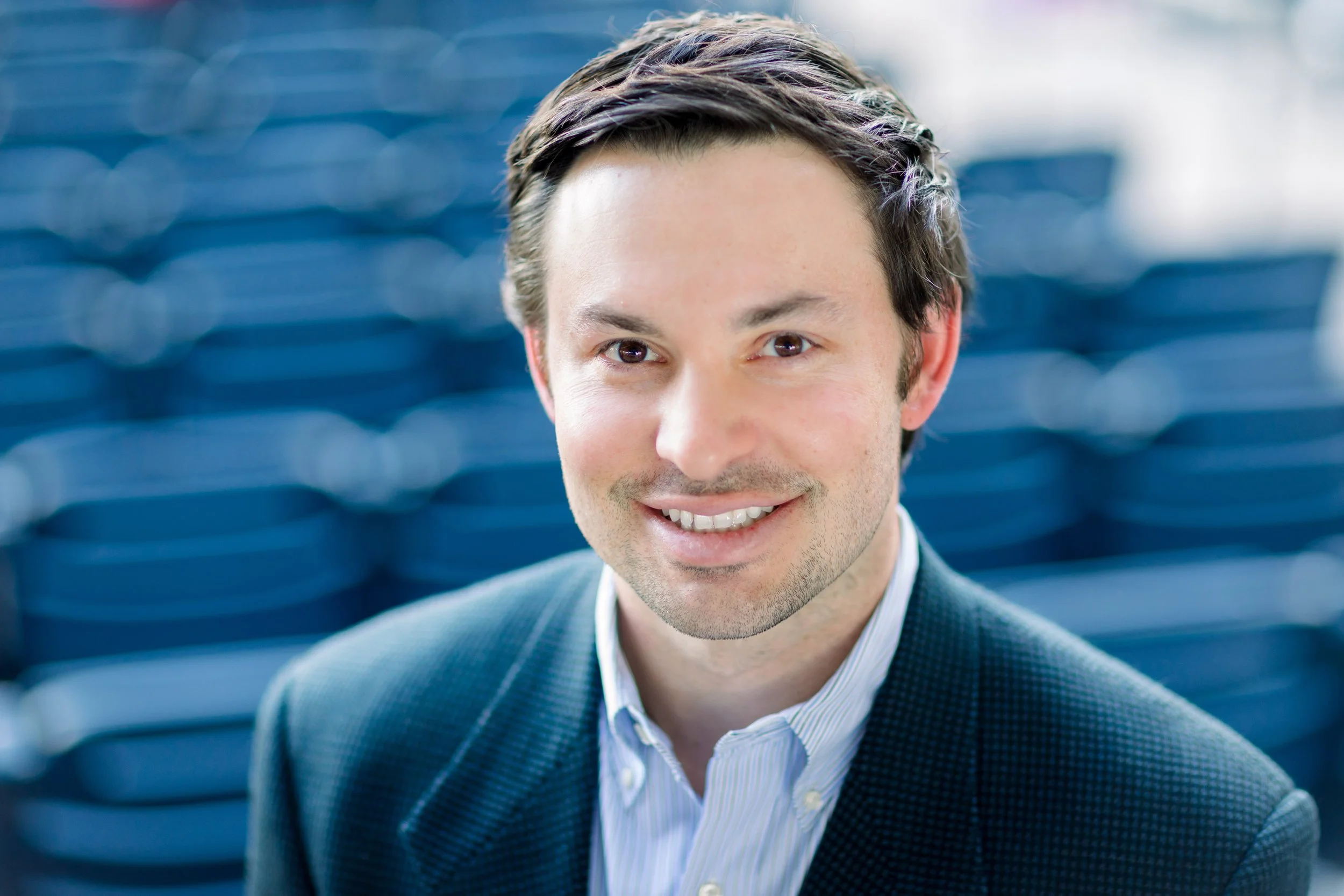 A young man dressed in a business suit smiling, with a blue stadium or arena seating background.