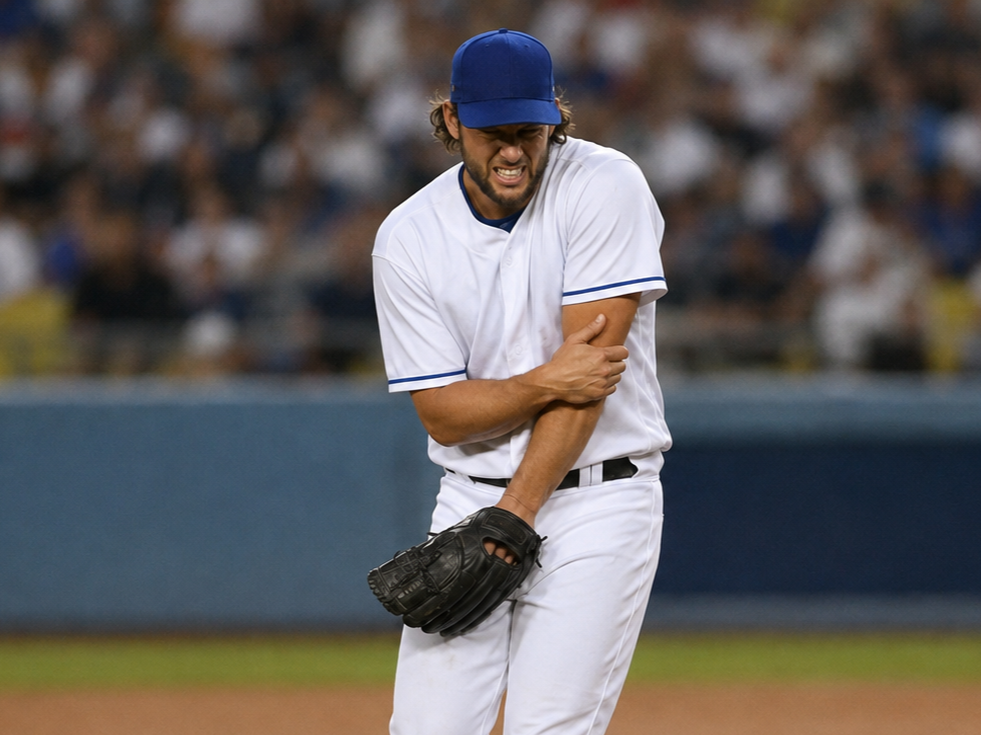 Baseball player grimacing and holding his arm in pain on the field, dressed in a white uniform and blue cap.