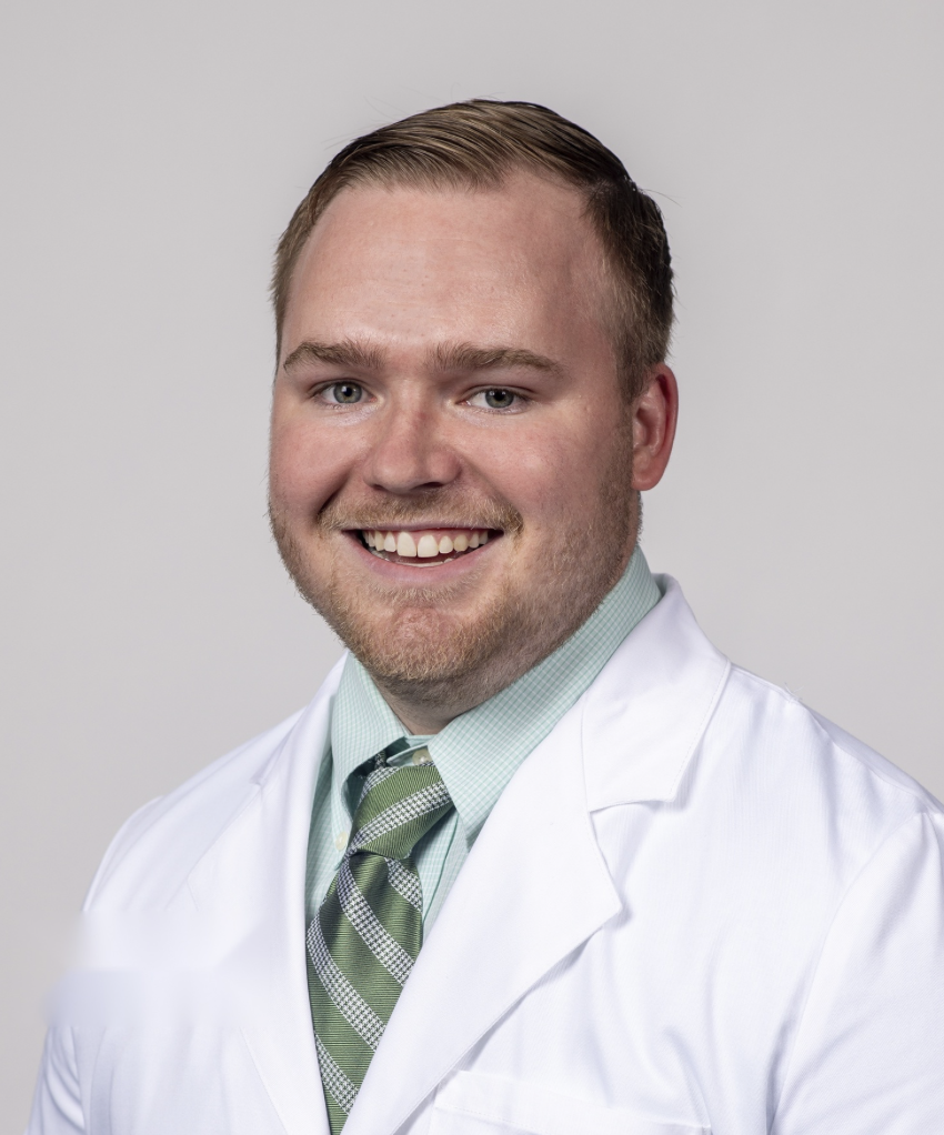 A smiling man in a white lab coat, light green dress shirt, and striped tie, posing against a plain light gray background.