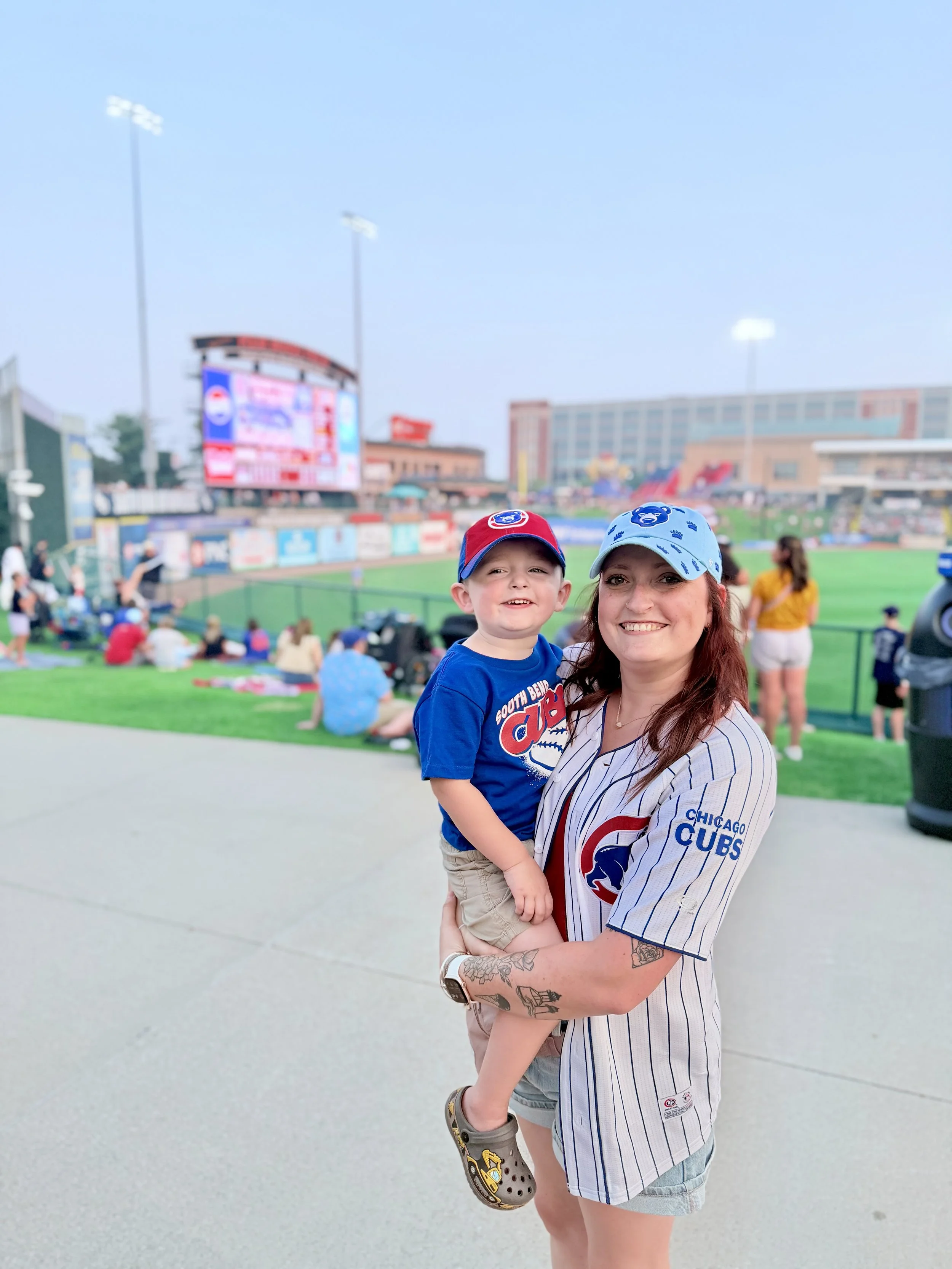 A woman with tattoos on her arms holding a young boy at a baseball stadium, both wearing Chicago Cubs gear, with the stadium crowd and scoreboard in the background.