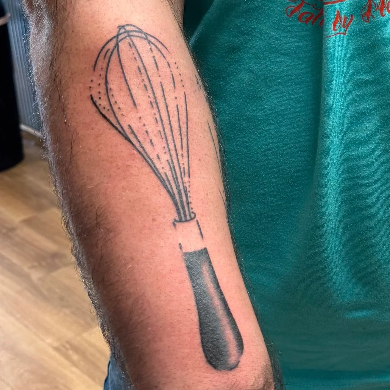 Tattoo of a stylized whisk with an outline and dotted shading on a person's upper arm.