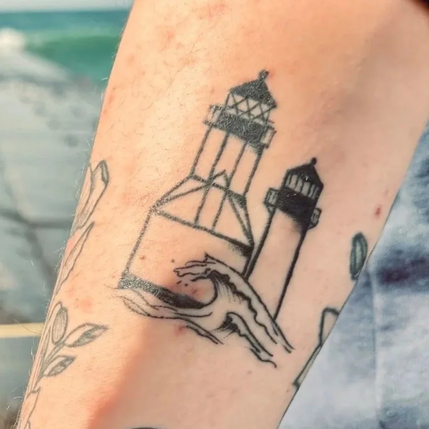 Tattoo of two lighthouses near the coast on a person's arm.