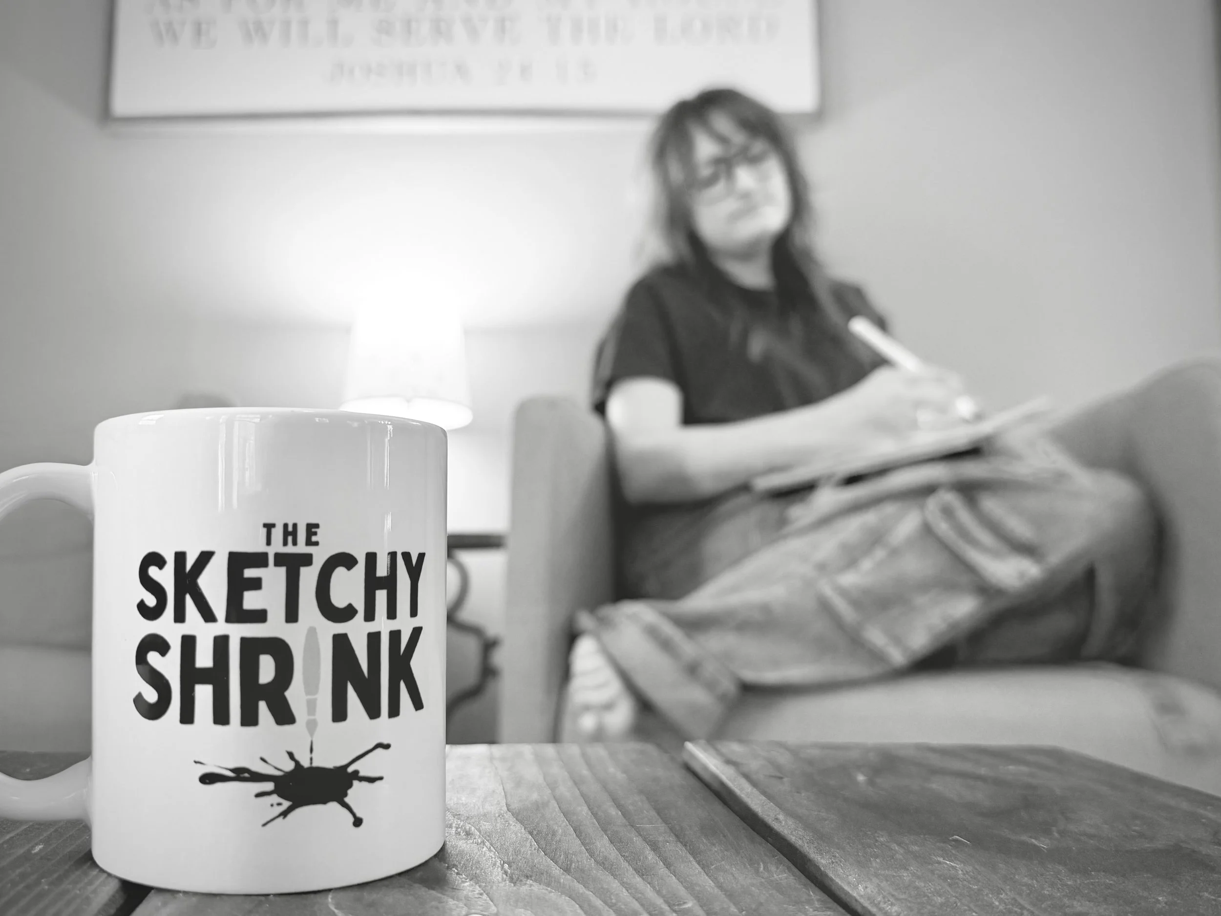 A white coffee mug with black text and a graphic of a bug on it in the foreground. Blurred person with glasses and layered hair sitting at a desk in the background, writing in a notebook, with a lamp on the desk and a framed quote on the wall behind them.