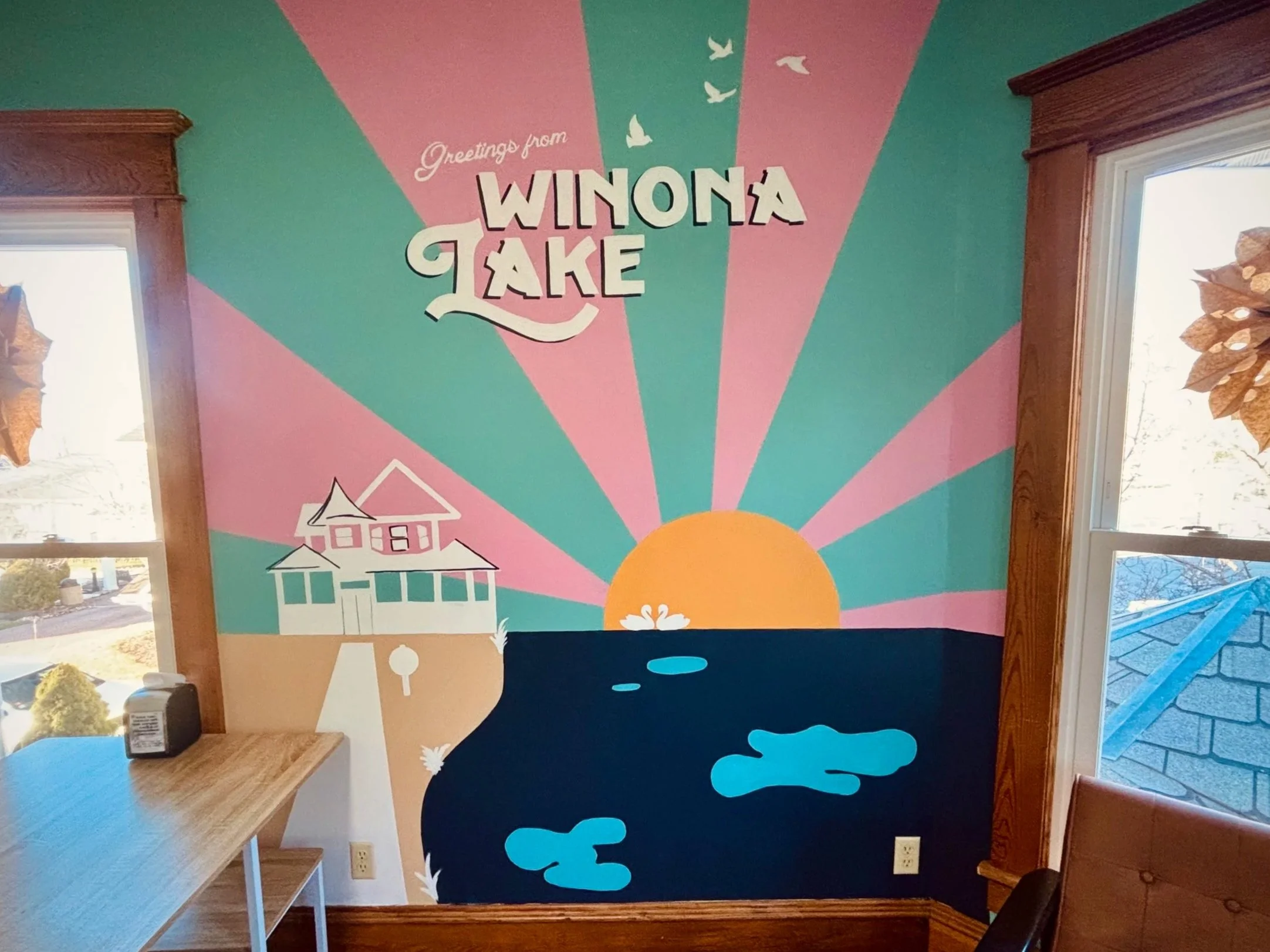 Colorful mural with pink and green rays, white birds, a house, and a pond or lake, with the phrase 'Greetings from Winona Lake' written on it.