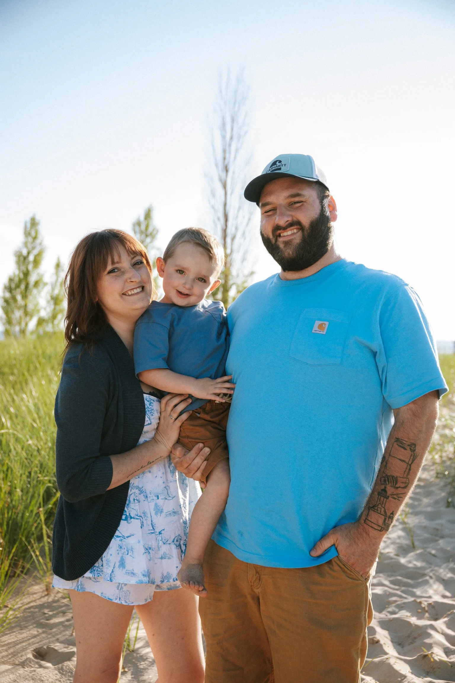 A happy family of three standing on a sandy beach with tall grasses and trees in the background, smiling at the camera on a sunny day.