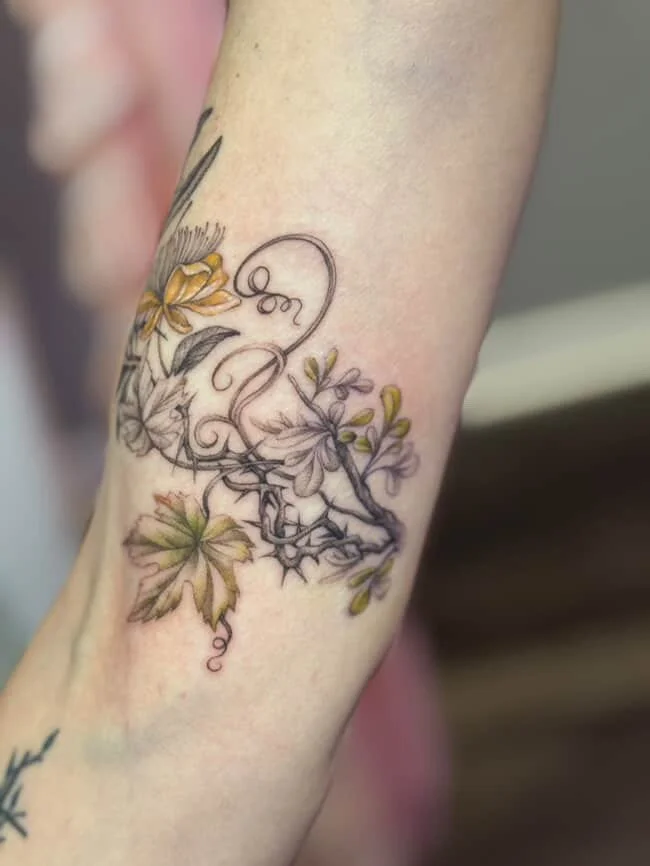 A tattoo on a person's forearm featuring a black and gray design of intertwined elements, with colored leaves and flowers in yellow, purple, and green.