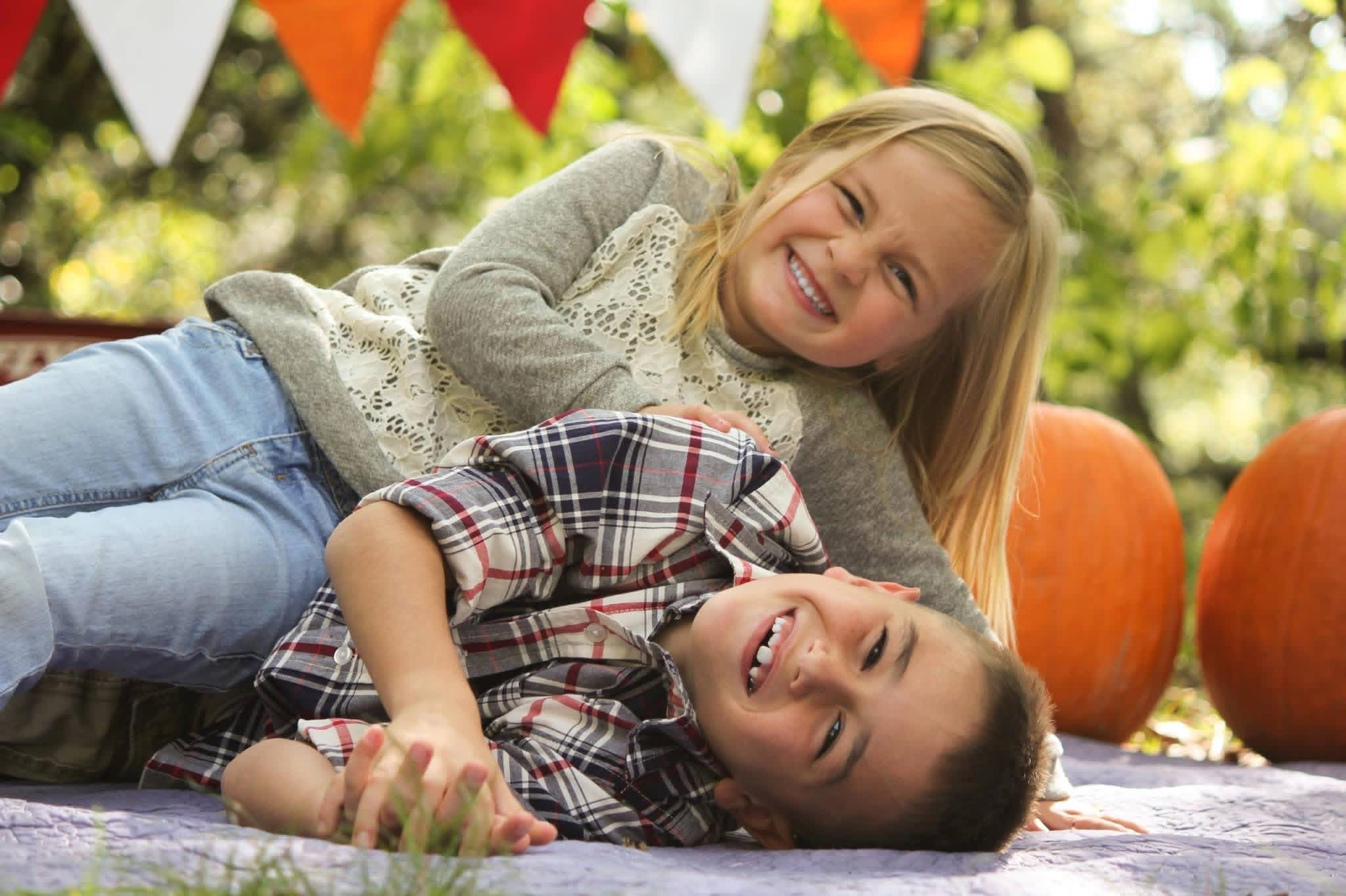 Two children lying on a blanket in an outdoor setting, smiling and playing, with pumpkins and colorful bunting in the background.