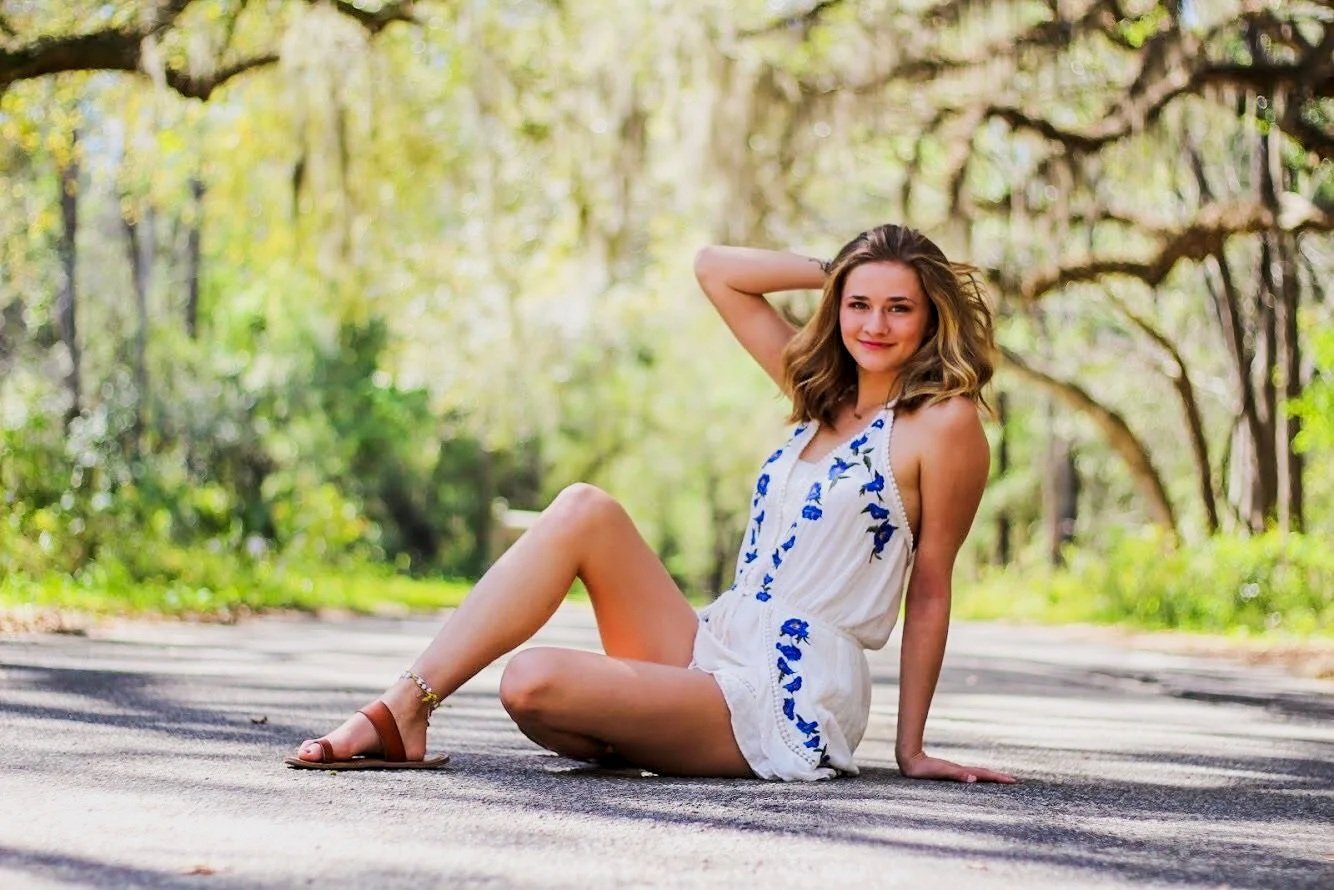 A woman sitting on a paved road in a sunlit forest, wearing a white dress with blue floral embroidery and brown sandals, smiling and looking at the camera.