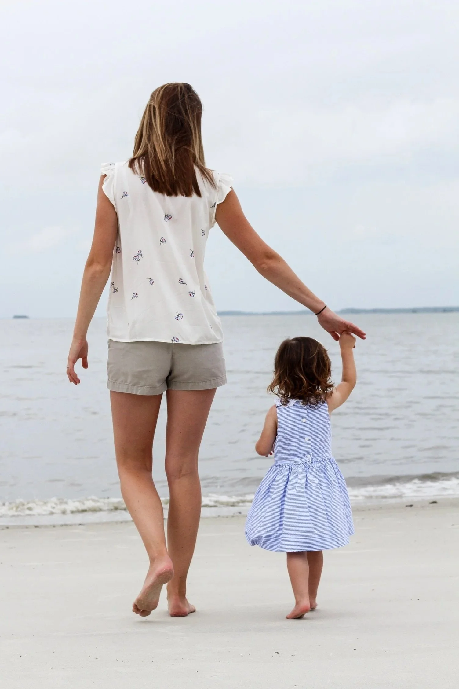 A woman and a young girl walking hand in hand on the beach, with the ocean and cloudy sky in the background.