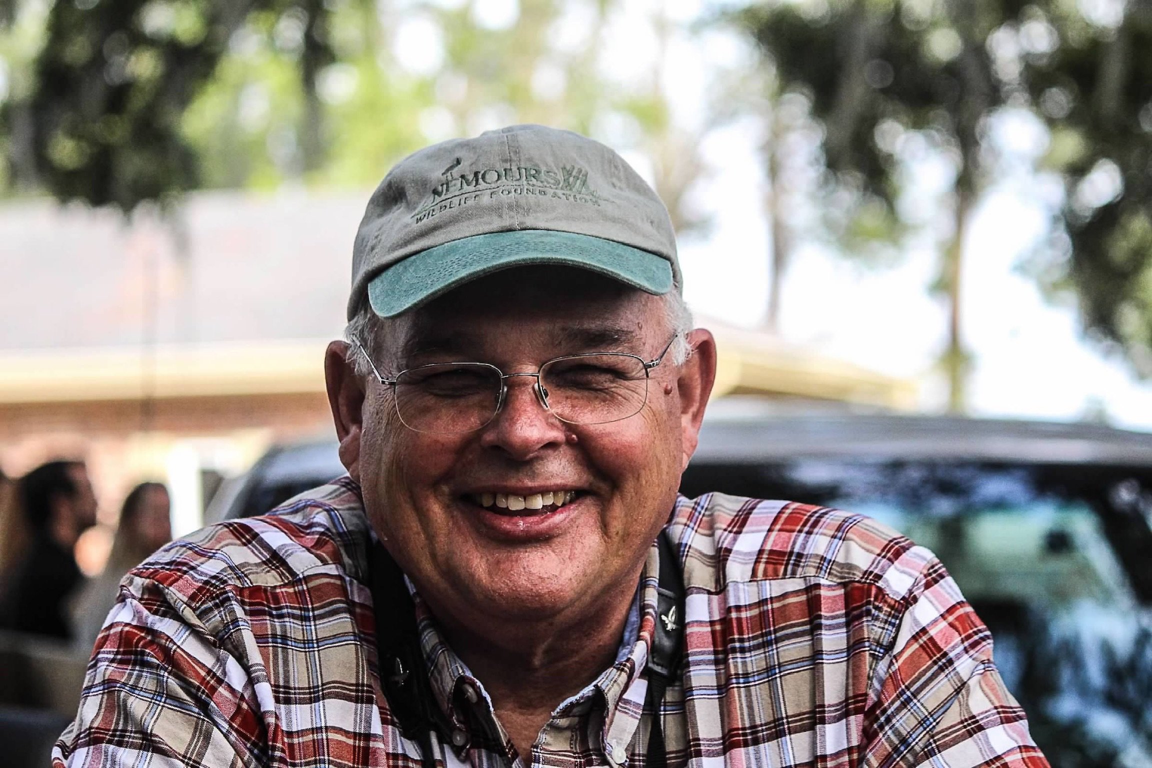 Close-up of smiling man wearing glasses, a gray cap, and a plaid shirt, outdoors with blurred trees and people in the background.