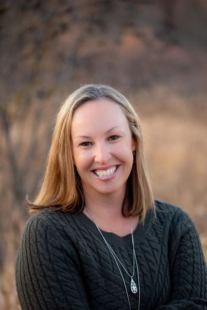 A woman with shoulder-length light brown hair smiling outdoors, wearing a dark knitted sweater and a pendant necklace.