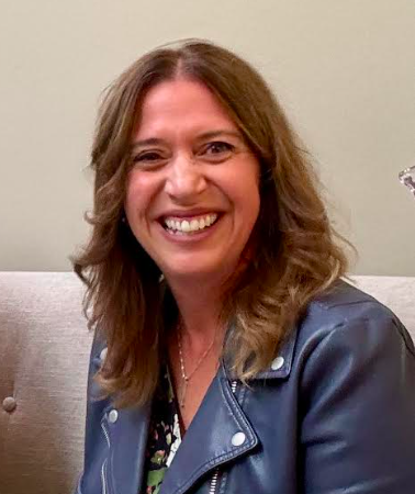 A woman with shoulder-length brown hair smiling, wearing a blue jacket, sitting indoors against a light-colored wall.
