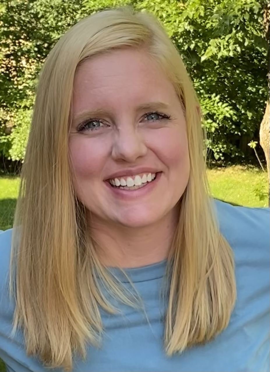A woman with long blonde hair, blue eyes, smiling, outdoors with green trees and sunlight in the background.