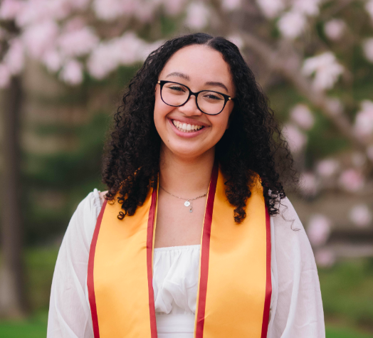A young woman with curly hair, glasses, and a white dress wearing a graduation stole, standing outdoors with blooming trees in the background.