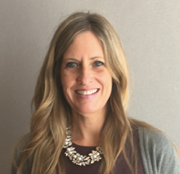 A woman with long blonde hair smiling, wearing a necklace and a gray blazer in front of a neutral background.