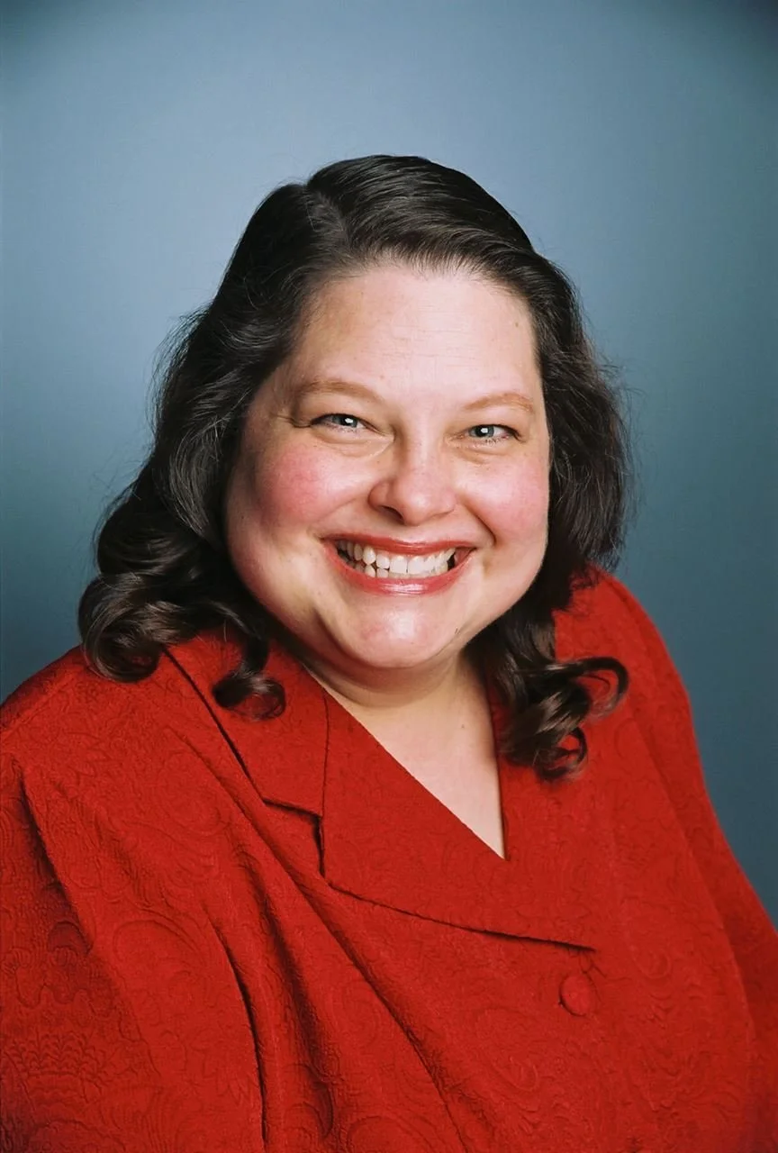 A smiling woman with dark, curly hair wearing a red top against a plain blue background.