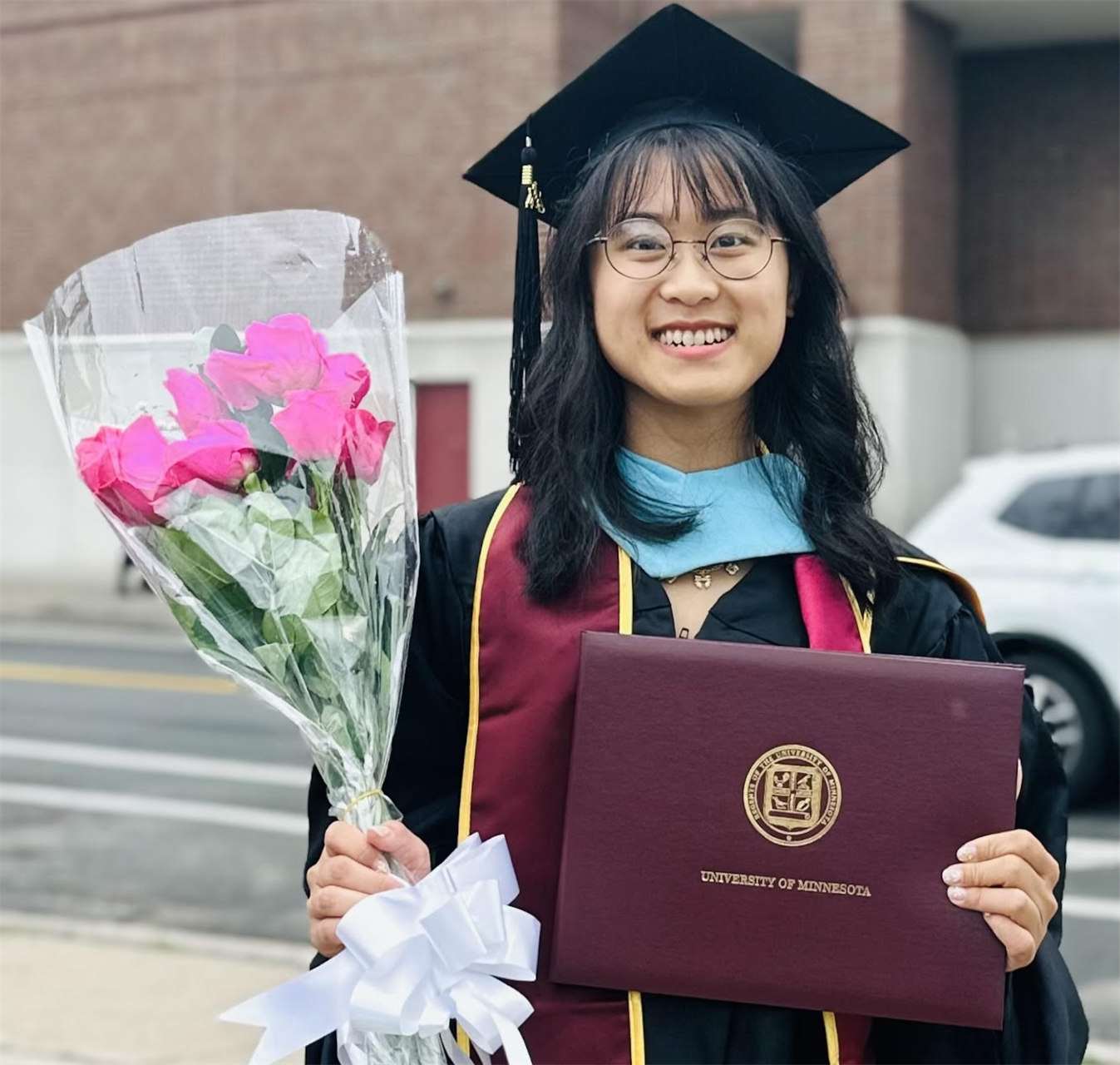 A young woman in graduation attire holding a bouquet of pink roses and a diploma from the University of Minnesota, smiling outside on a street.