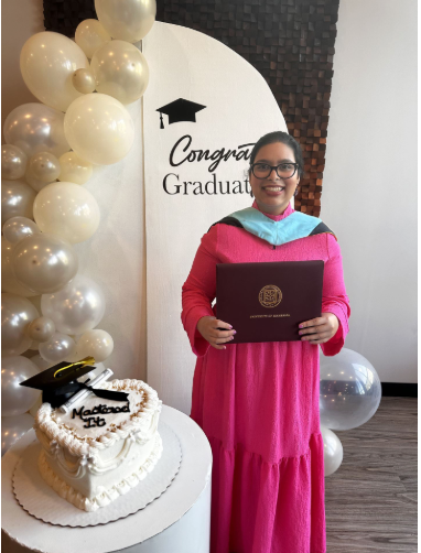 A woman in a bright pink gown and glasses holding a diploma, standing next to a graduation cake with a black mortarboard decoration. The background has a white sign with a graduation cap icon and the words 'Congrats Graduate,' along with gold and white balloons.