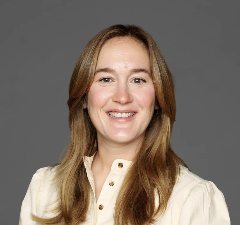 A woman with long, light brown hair smiling at the camera, wearing a cream-colored blouse with gold buttons, against a gray background.