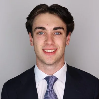 Portrait of a young man with dark hair, blue eyes, wearing a suit and tie, smiling against a neutral background.
