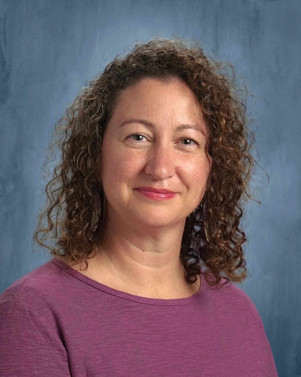 A woman with curly brown hair and light skin, wearing a purple top, smiling against a blue textured background.