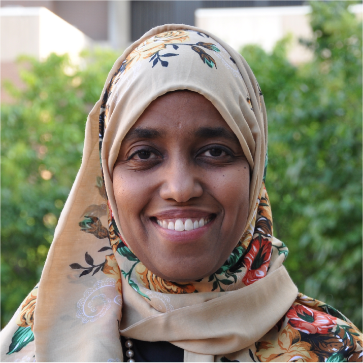 A woman wearing a beige floral-patterned headscarf, smiling outdoors with greenery in the background.