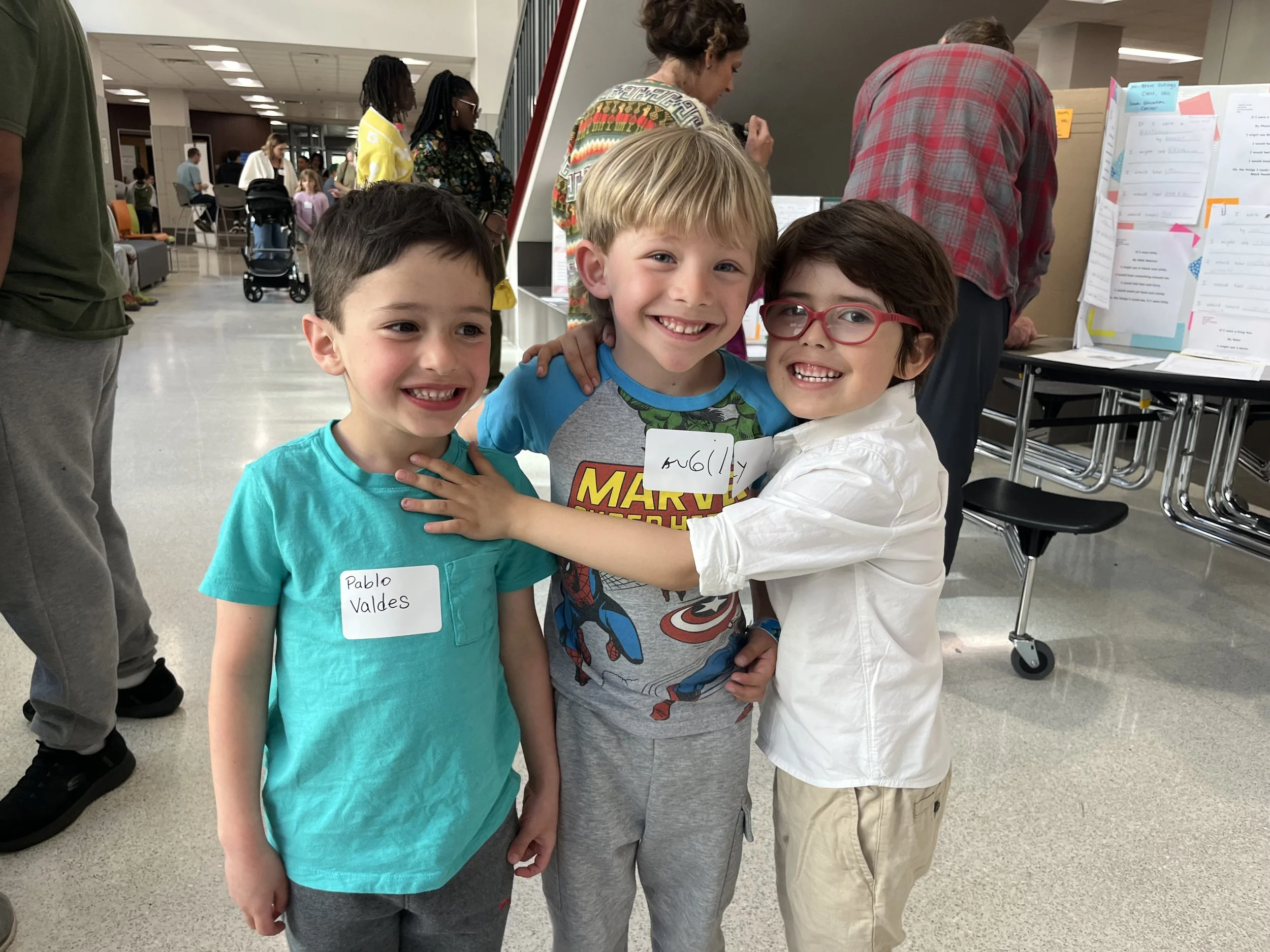 Three young boys standing close together, smiling and hugging indoors at an event. The boy on the left has short dark hair, the middle boy has blonde hair and is wearing a Marvel-themed shirt, and the boy on the right has dark hair, red glasses, and is wearing a white shirt.