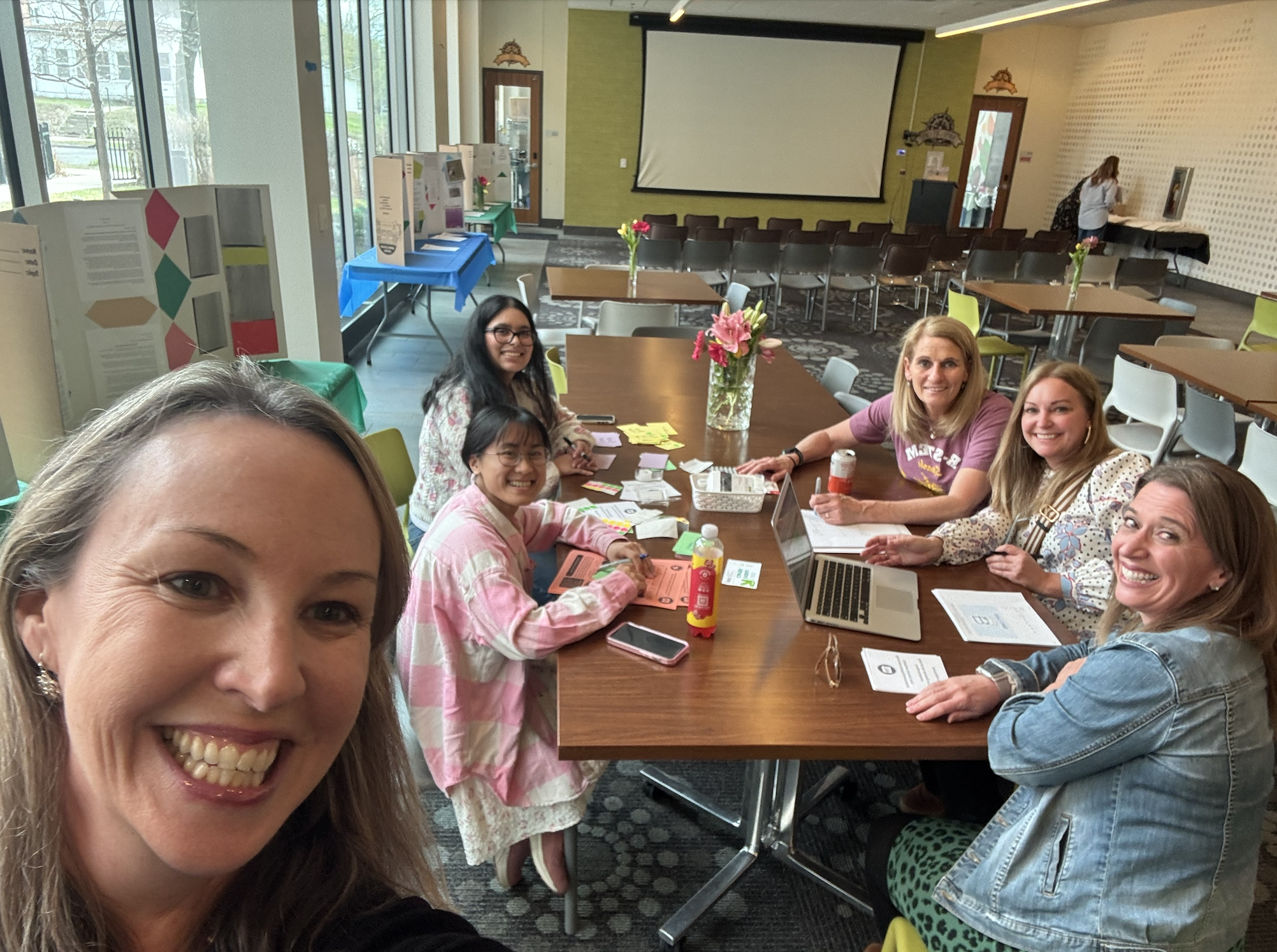 Group of women smiling at a table during a meeting or workshop in a room with large windows, presentation screen, and additional tables with informational displays.