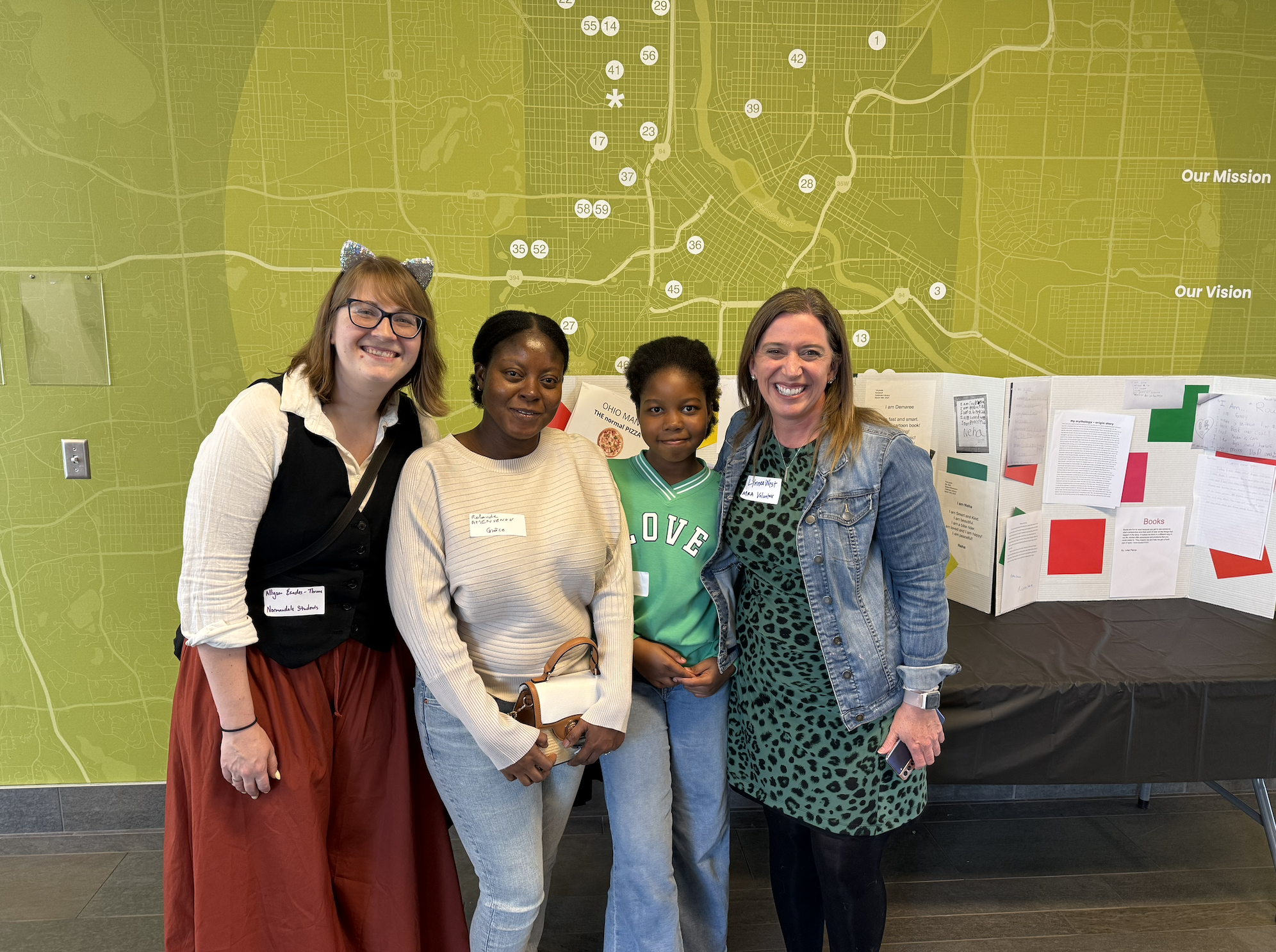 Four women standing together indoors in front of a large map display, smiling at the camera. One woman is wearing a cat ear headband, glasses, and a black and white outfit; another has a beige sweater and a name tag; the third is a young girl in a green sports jersey; and the fourth woman is in a blue denim jacket with a badge and patterned dress.