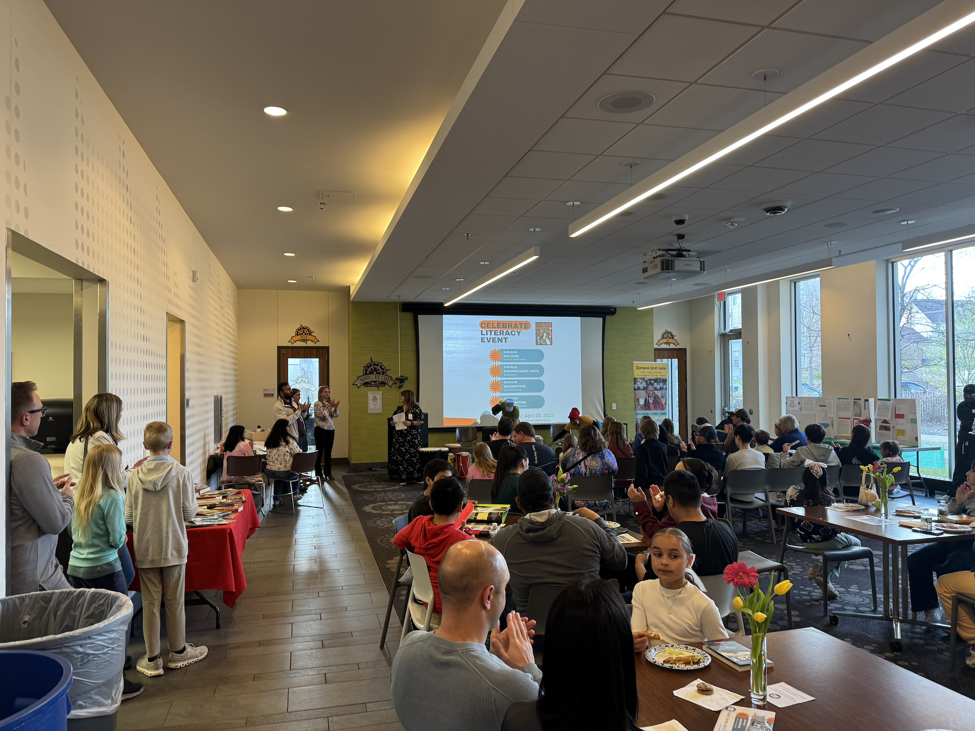 A large room filled with people sitting at tables with snacks and flowers, watching a presentation on a screen that reads 'Celebrate Literacy Event' with a schedule and artwork. The room has large windows and ceiling lights.