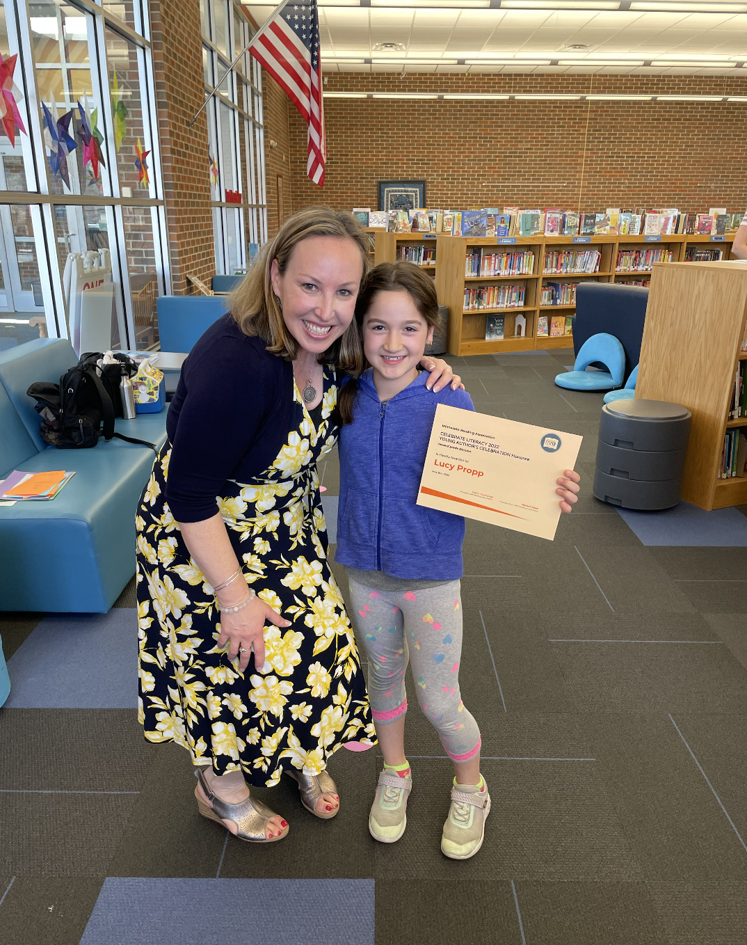 A woman and a young girl smiling and posing together inside a library, with the girl holding a certificate of achievement. Bookshelves and an American flag are visible in the background.