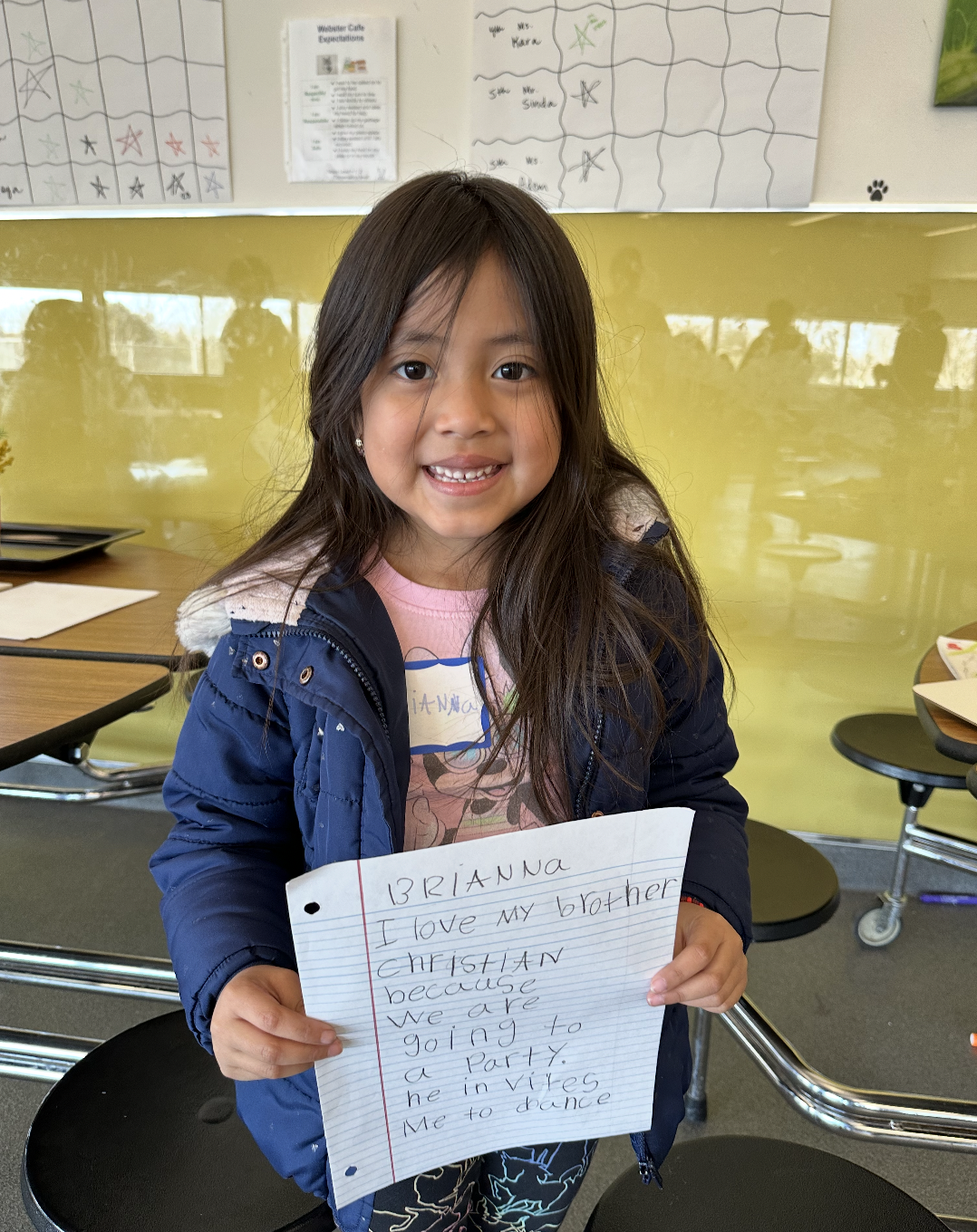 A young girl with long dark hair holding a handwritten note, smiling in a classroom with yellow and white walls and charts in the background.
