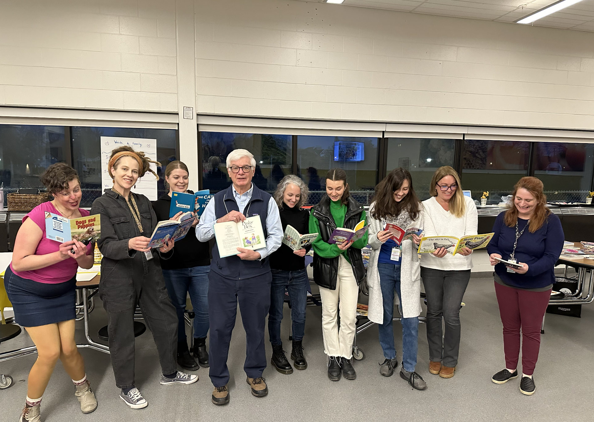A group of nine people standing together indoors, holding open books, smiling, and appearing engaged in reading or discussing. The setting looks like a community center or classroom with windows in the background and tables with various items.