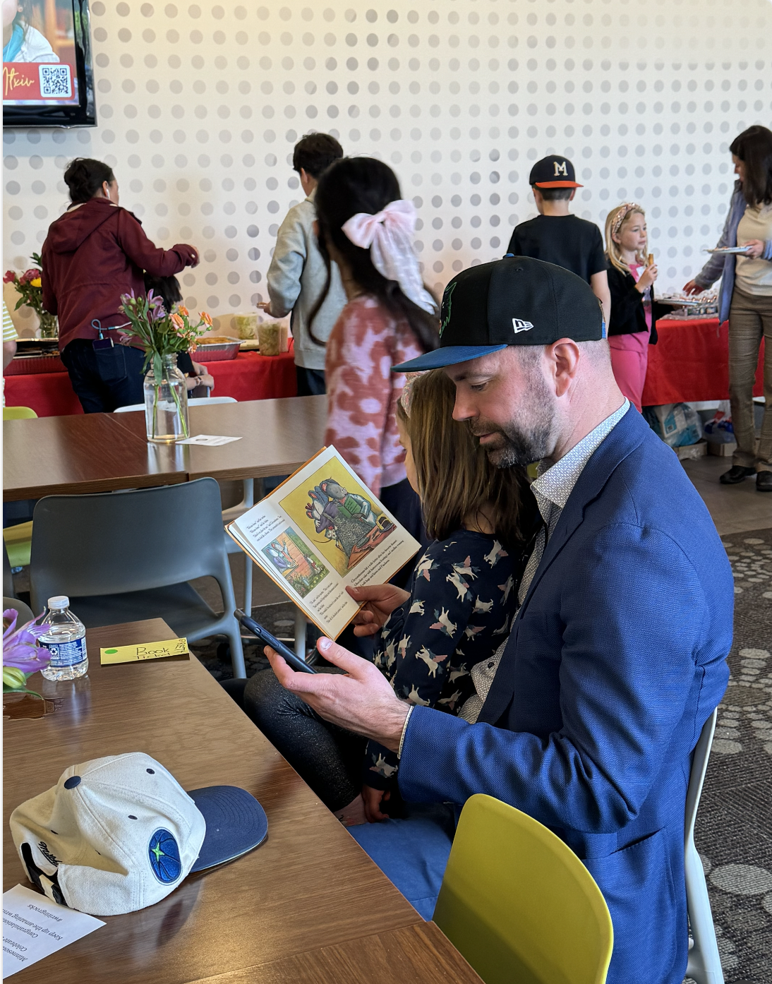 A man in a blue suit and black cap sitting at a table, reading a children's book, with a girl sitting next to him. On the table are a water bottle, a beige cap, and a yellow note. In the background, a group of children and women are in a room with dotted wallpaper and a red tablecloth, some serving food.