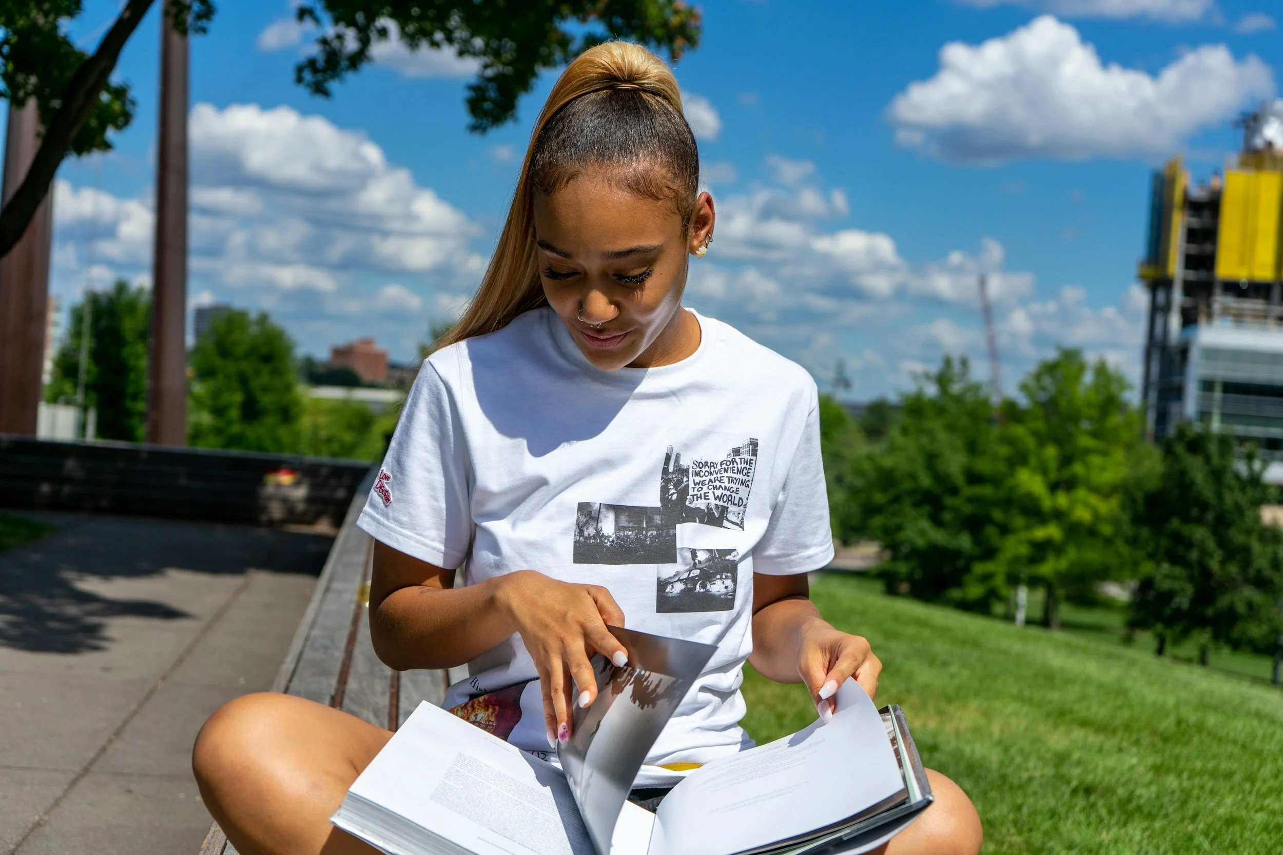 A young woman sitting outdoors on a bench, looking down at and flipping through papers, with green trees and buildings in the background, under a partly cloudy sky.