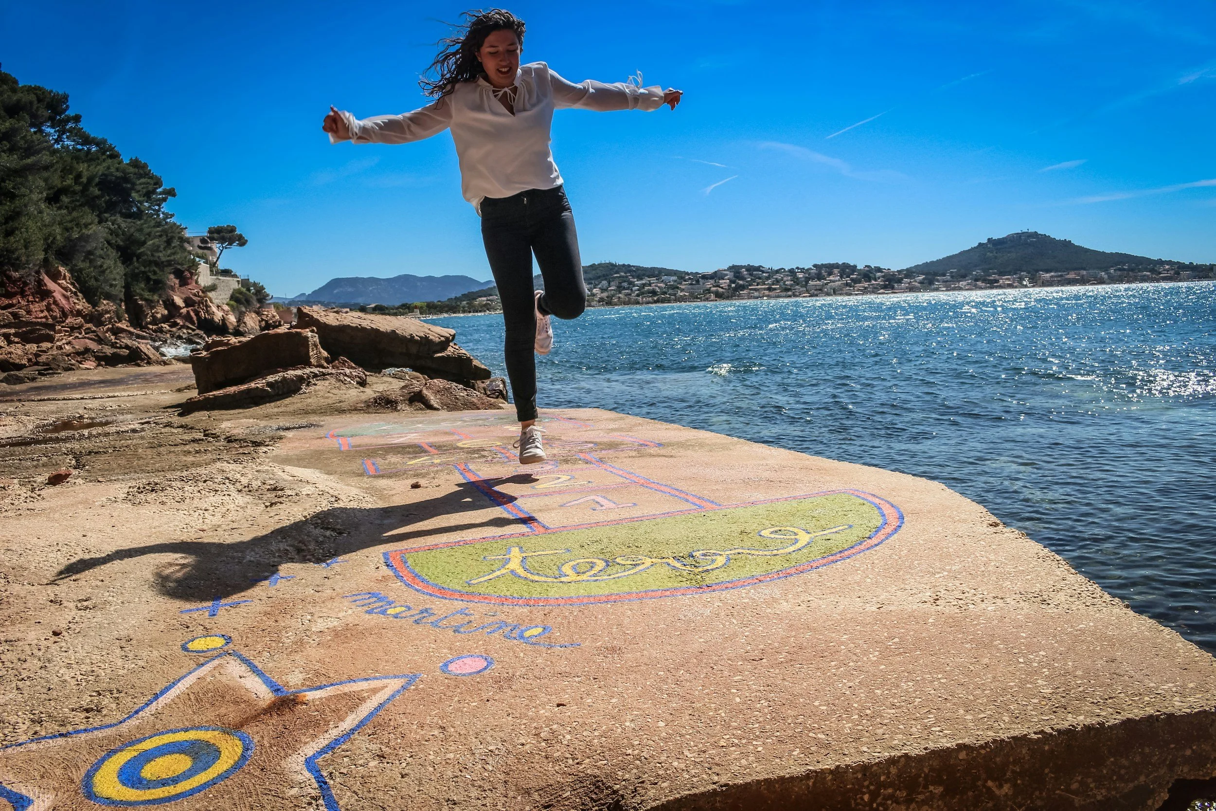 A woman jumping on a hopscotch court near the water with a mountainous landscape in the background under a clear blue sky.