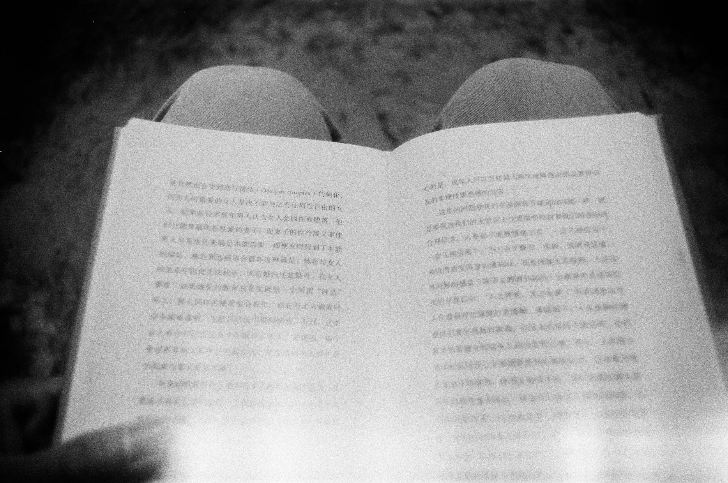 Black and white photo of an open book with Chinese characters, viewed from above, with legs in jeans visible in the background.