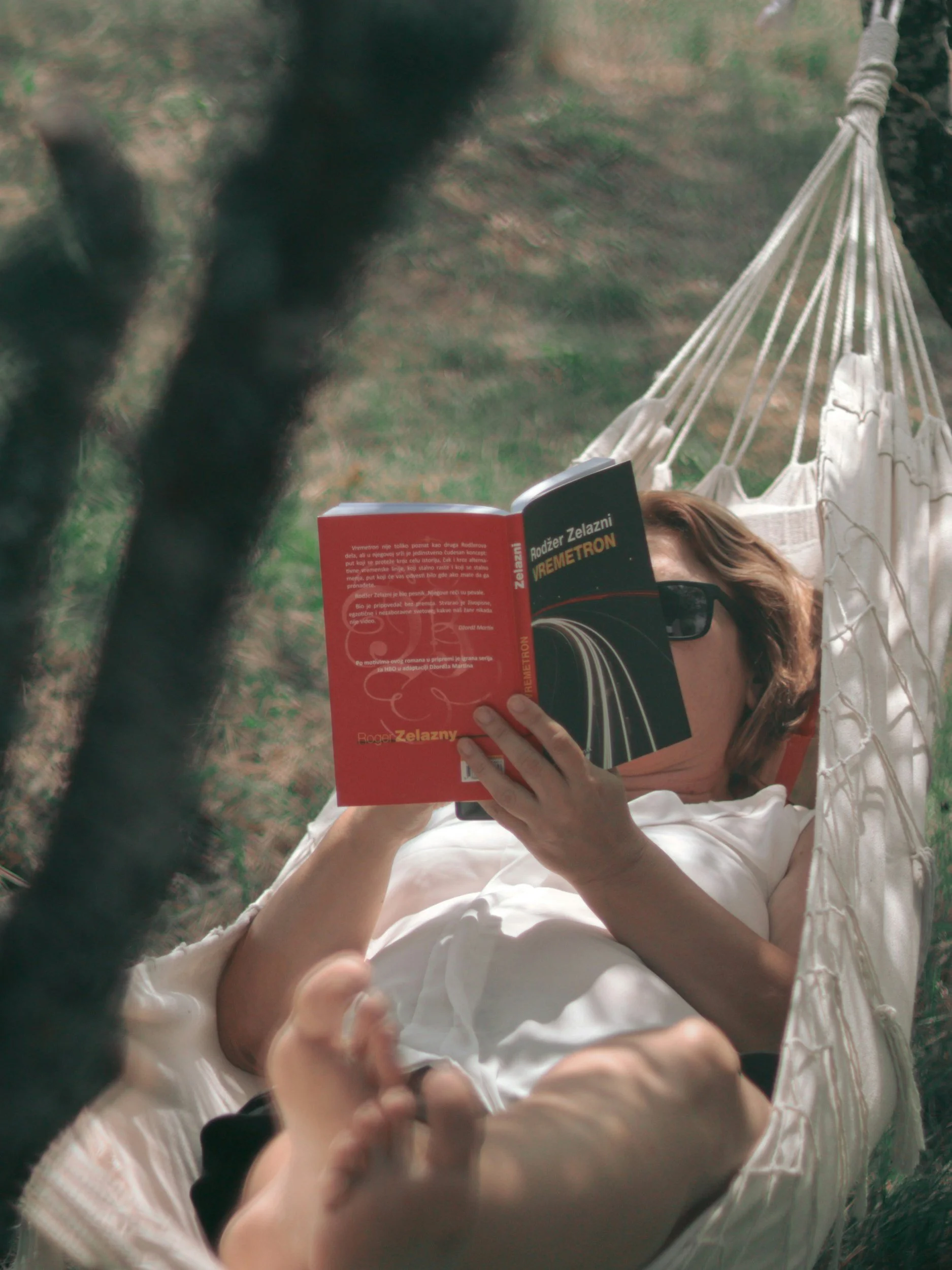 A person is lying in a hammock outdoors, reading a book titled 'Rodz Zelażny REMETRON'. The person has glasses and brown hair. The hammock is tied to trees, and the setting appears to be in a grassy area with some trees in the background.