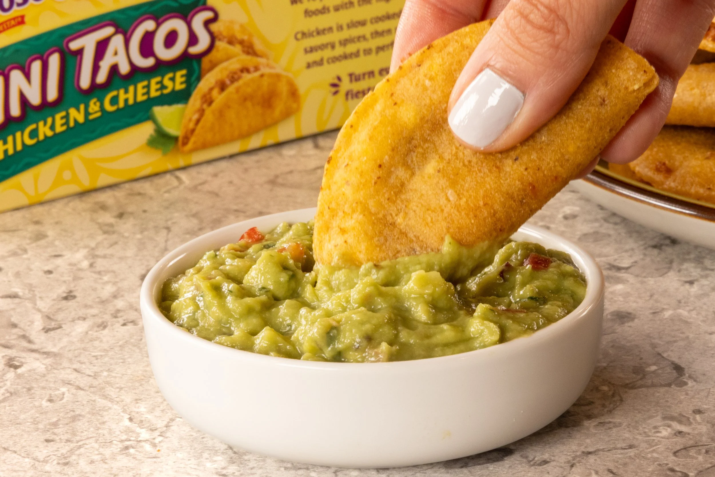 A hand with white painted fingernails holding a fried taco with guacamole in a white bowl in the foreground. In the background is a yellow box of chicken tacos and a plate of more tacos.