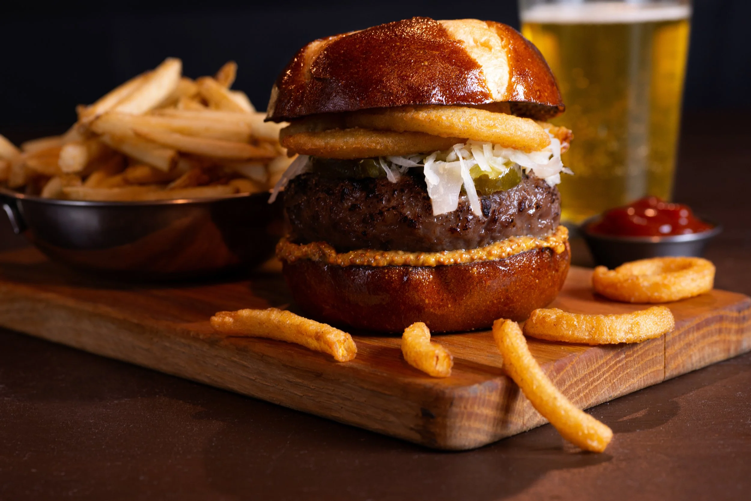 A juicy burger with a sesame seed bun topped with pickles, coleslaw, onion rings, and mustard, served on a wooden board, alongside a cup of French fries, ketchup, and a glass of beer.