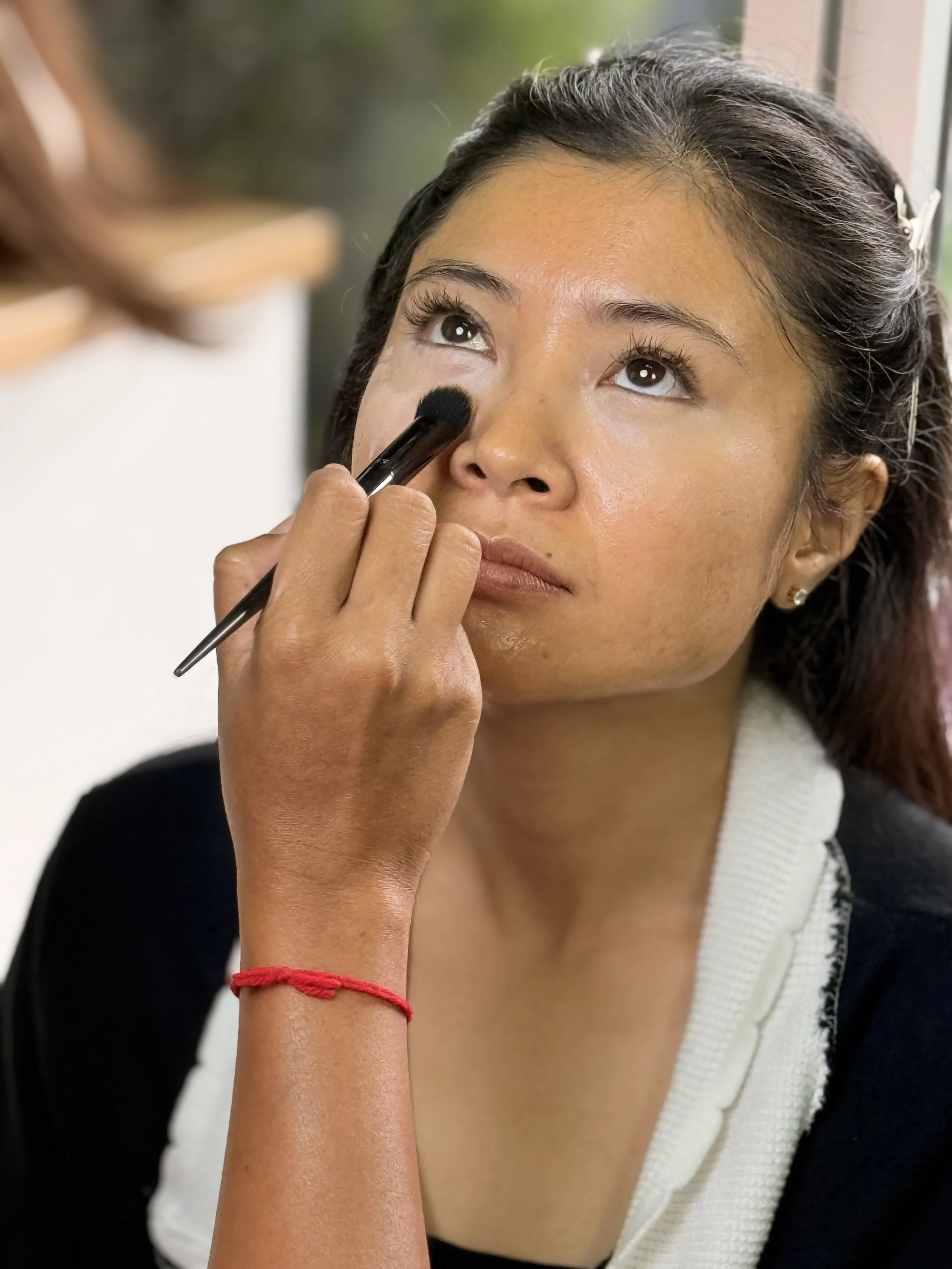 A woman with dark hair and light brown skin gets her makeup done, with a makeup brush near her eye.